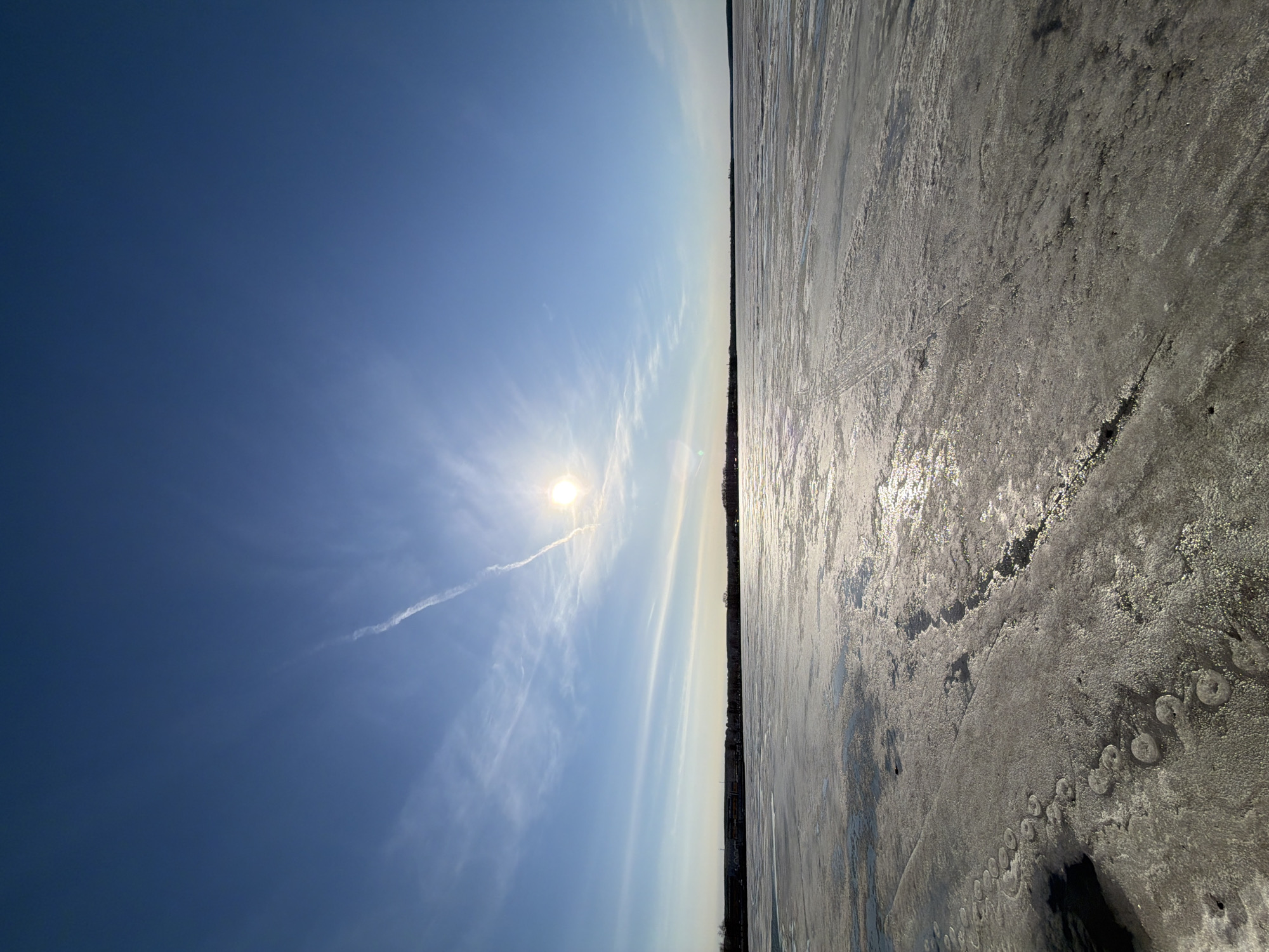 The image shows a frozen body of water, possibly a lake or wide river, bisected by a distant treeline. The ice surface is textured and appears to be melting, reflecting the sun. A portion of the sky is visible, with a clear blue color and wispy clouds. The sun is bright and surrounded by a diffuse glow. A contrail is also visible. The location, according to the prompt, is in Votkinsk, Russia. Details about the city and its specific geographical features may help identify the body of water depicted.