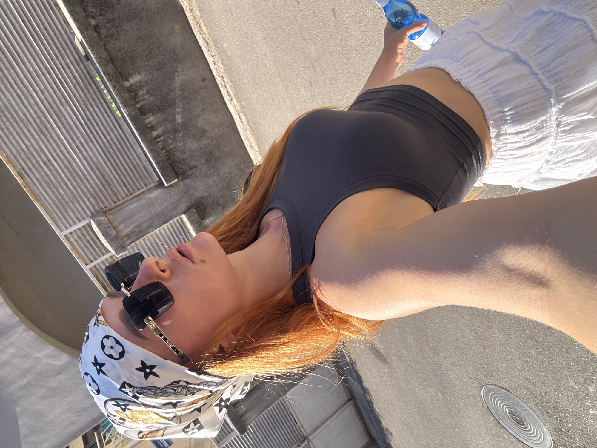 In Budva, Montenegro, a young woman with long, bright orange hair is pictured from the chest up, walking outdoors on a paved street. She is wearing a white bandana with a black and gold pattern, large black sunglasses, a dark grey cropped tank top, and a white tiered ruffled skirt. She is holding a blue water bottle in her right hand. The background shows a concrete wall and a ribbed metal structure.