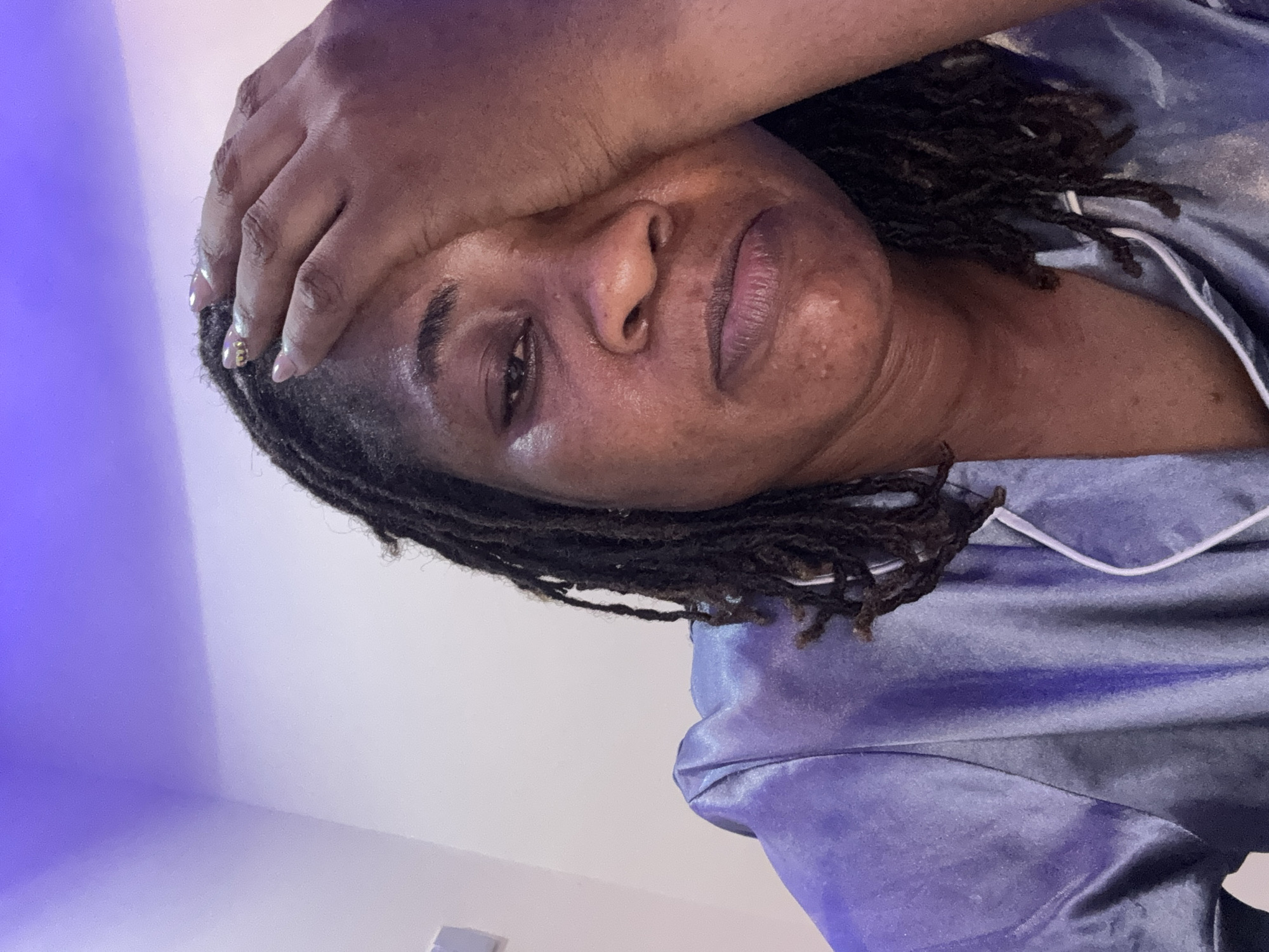 The image is a close-up, selfie-style portrait of a Black woman with dreadlocks. She is wearing a light blue, satin pajama top with white piping. Her right hand is resting on her forehead and temple, partially obscuring her right eye. Her expression appears weary or distressed, with her mouth slightly downturned. The background consists of a wall with a purple light cast on the upper portion, suggesting an artificial light source, possibly from an LED strip. The woman's skin shows some texture and minor blemishes. There are no specific landmarks or environmental cues that would indicate a location in Ajara, Nigeria. The scene is indoors, and the focus is on the woman's face and upper body.
