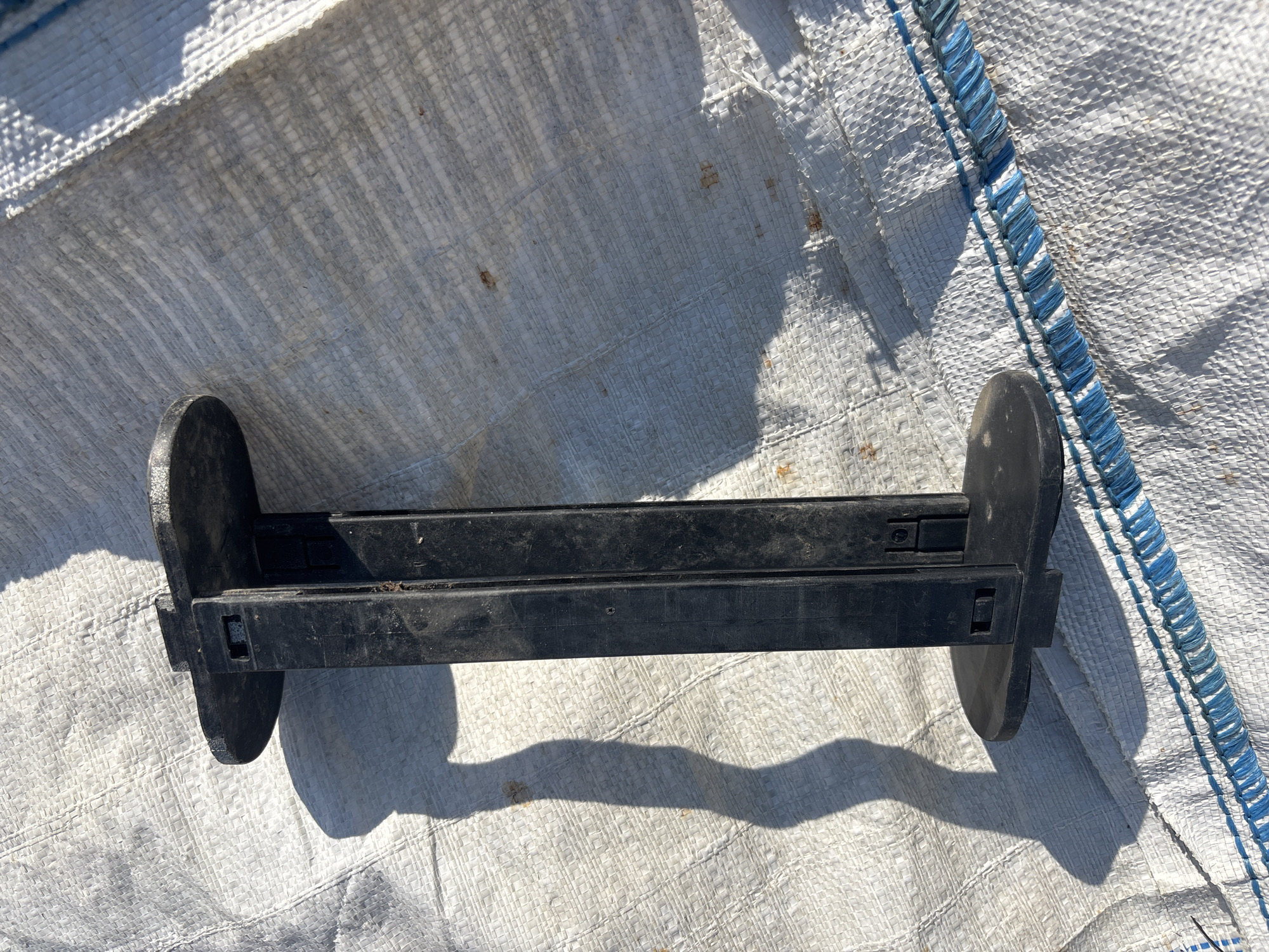 A black plastic object, resembling a spool or a tool, rests on a white woven tarp. The object has two flat, circular ends connected by a rectangular central piece. It casts a long, distorted shadow to the left, suggesting it is outdoors under direct sunlight. The tarp is textured and shows some reddish-brown stains. Blue stitching runs along the bottom edge of the tarp. The scene is situated in Arkhipovskoe, Russia.