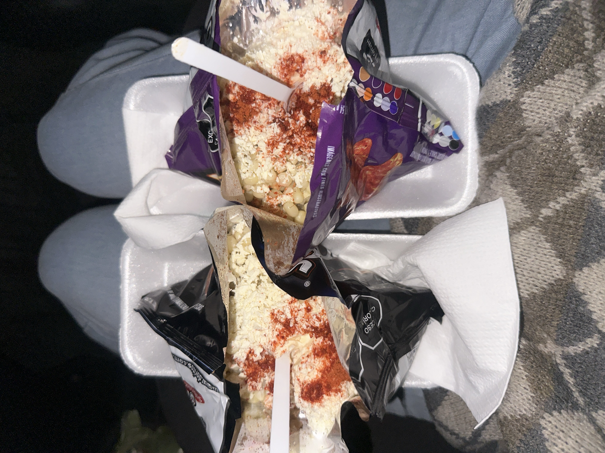 Here is a description of the image:
The image shows two servings of "elotes" or Mexican street corn. Each serving is in a clear plastic bag, placed in a white styrofoam container. The elote is topped with white cheese and a generous dusting of red chili powder. A white plastic spoon is stuck into each serving. The elotes are sitting on a person's lap, one serving on each thigh. The person is wearing a light blue pant leg on the top half of the photo and a grey and brown patterned sweater on the bottom half of the photo. A partially visible black surface is on the left side of the frame. Some of the packaging has the words "EXC", "CHILEM", and "www.doritos.com" in the upper right corner and "IMAGENES CON FINES ILUSTRATIVOS" in purple lettering, which suggests the dish may be flavored with Doritos. The city location is unknown, as is the setting.