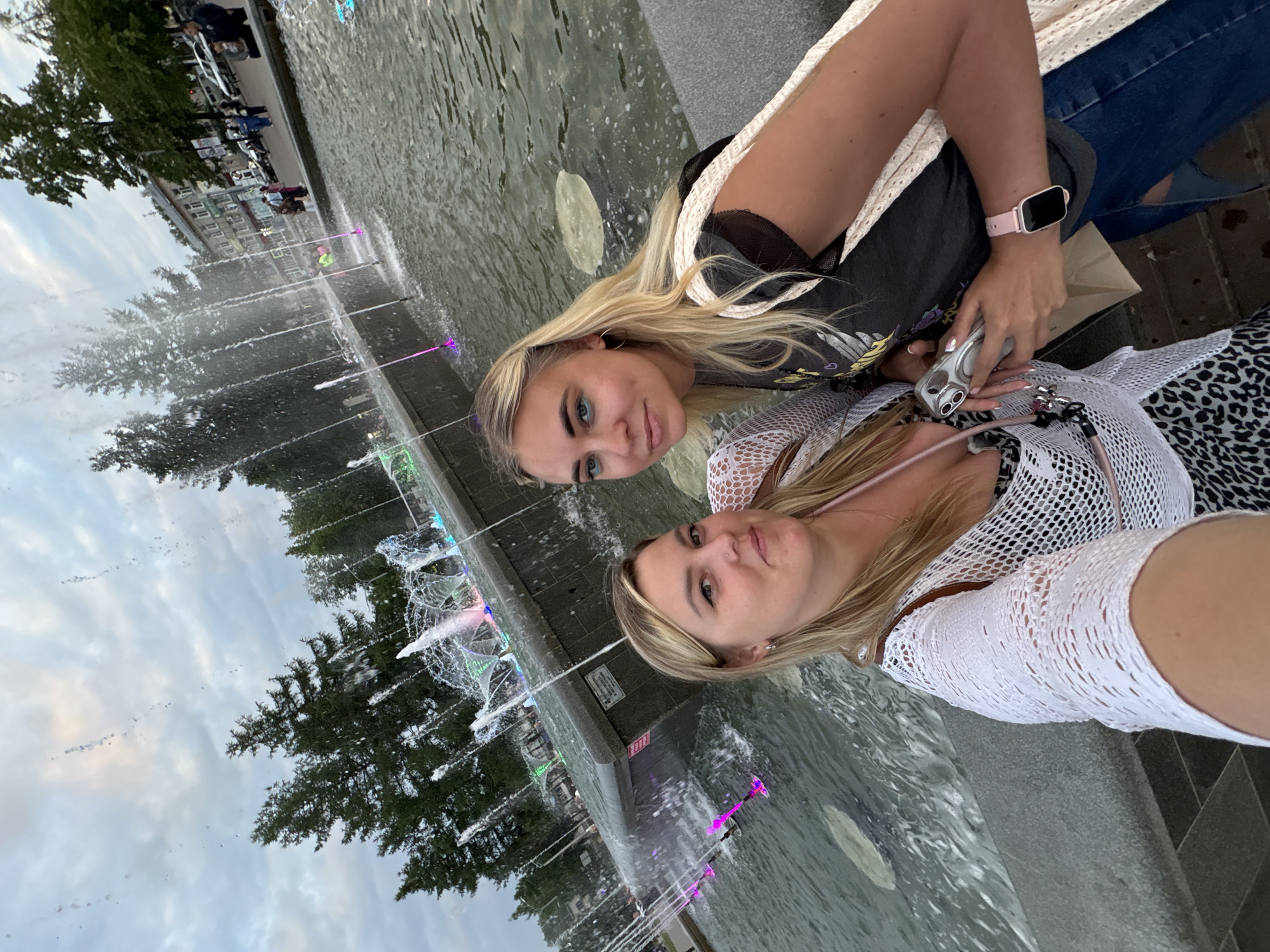 The image shows a selfie of two blonde women in front of a fountain. The woman on the left is wearing a white lace top and the woman on the right is wearing a dark t-shirt and jeans. Both women are smiling. The fountain is lit with colorful lights and is located in a city square. In the background, there are trees and buildings. This fountain is located in Chaikovsky, Russia.