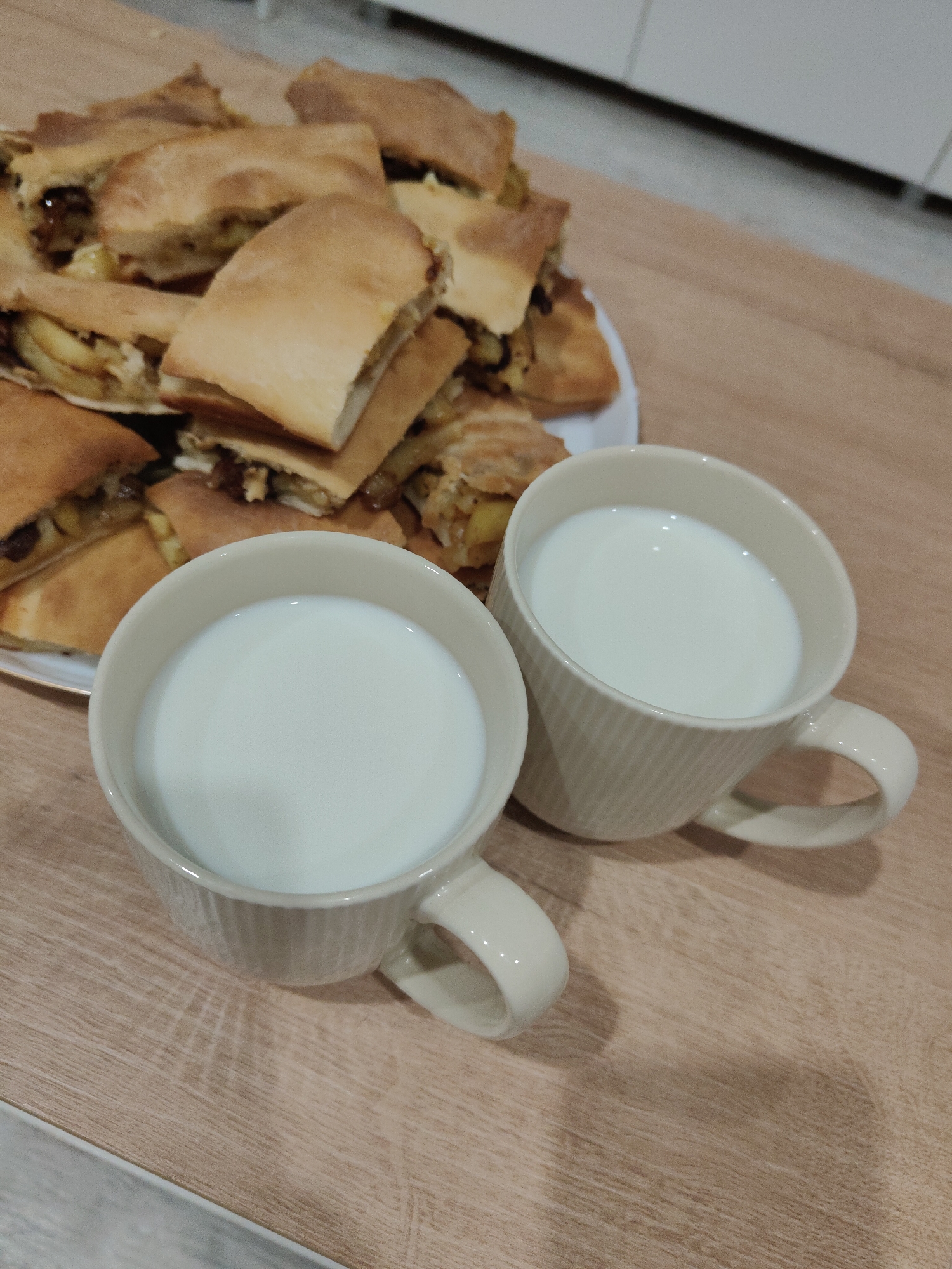 The image shows two mugs of milk and a plate of baked goods, likely pie or turnover, on a wooden table. The location is unknown.