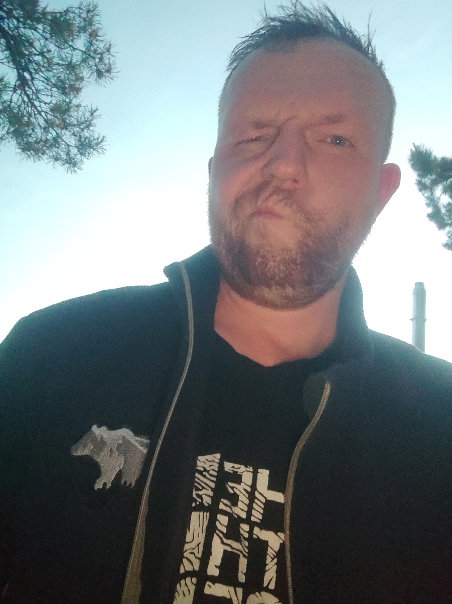 This is a waist-up selfie of a man outdoors, with the sky and trees in the background. The man has short, spiked blond hair, a beard, and is making a face as if he is squinting or surprised. He is wearing a dark jacket over a black t-shirt. On the left side of his jacket, there is a patch of a grey bear. On his t-shirt, there is a white graphic design. The background suggests it is daytime, with a clear sky and some branches from a tree on the left and right sides of the frame. There is a tall, cylindrical industrial structure visible in the distance on the right. The image was taken in Motornoe, Russia.