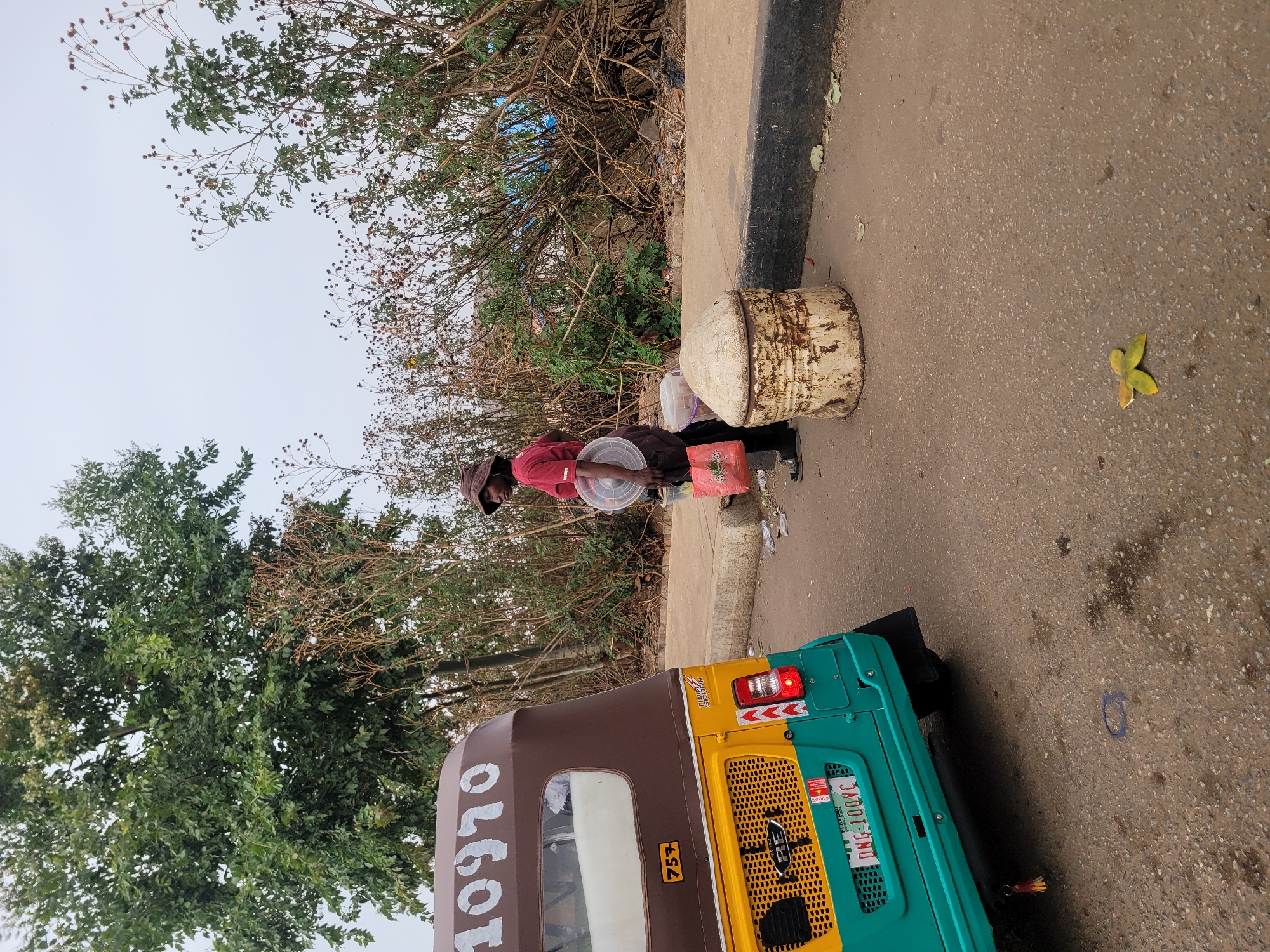 This daytime outdoor image, likely under an overcast sky, captures a street scene in Jos, Nigeria.
In the foreground, the rear section of a three-wheeled auto-rickshaw (keke napep) is visible on the left. Its canopy is brown with white numbers "10970" displayed. The vehicle features a yellow mid-section and a teal/green rear. A yellow sticker with "75+" is present, along with the brand name "TRISHAW". The visible license plate, "ONG-100XC," clearly states "PLATEAU" and "NIGERIA" below, identifying its registration.
To the right of the vehicle, a person, likely a street vendor, stands by the concrete curb on the paved asphalt road. They wear a dark head covering, a red long-sleeved shirt, dark trousers, and dark shoes. The vendor is holding multiple clear plastic containers, suggesting they are selling food or goods. A red plastic bag, featuring the "FIFA WORLD CUP" logo, rests on the ground near them. Adjacent to the vendor, a worn, light-colored barrel-like object sits on the curb.
The background is filled with varied vegetation, including trees with lush green leaves and bushes with drier, sparser branches and seed pods, extending towards a light gray, overcast sky. A single yellow-green leaf lies on the asphalt in the mid-ground. The scene depicts typical roadside commerce in a Nigerian urban or semi-urban environment.