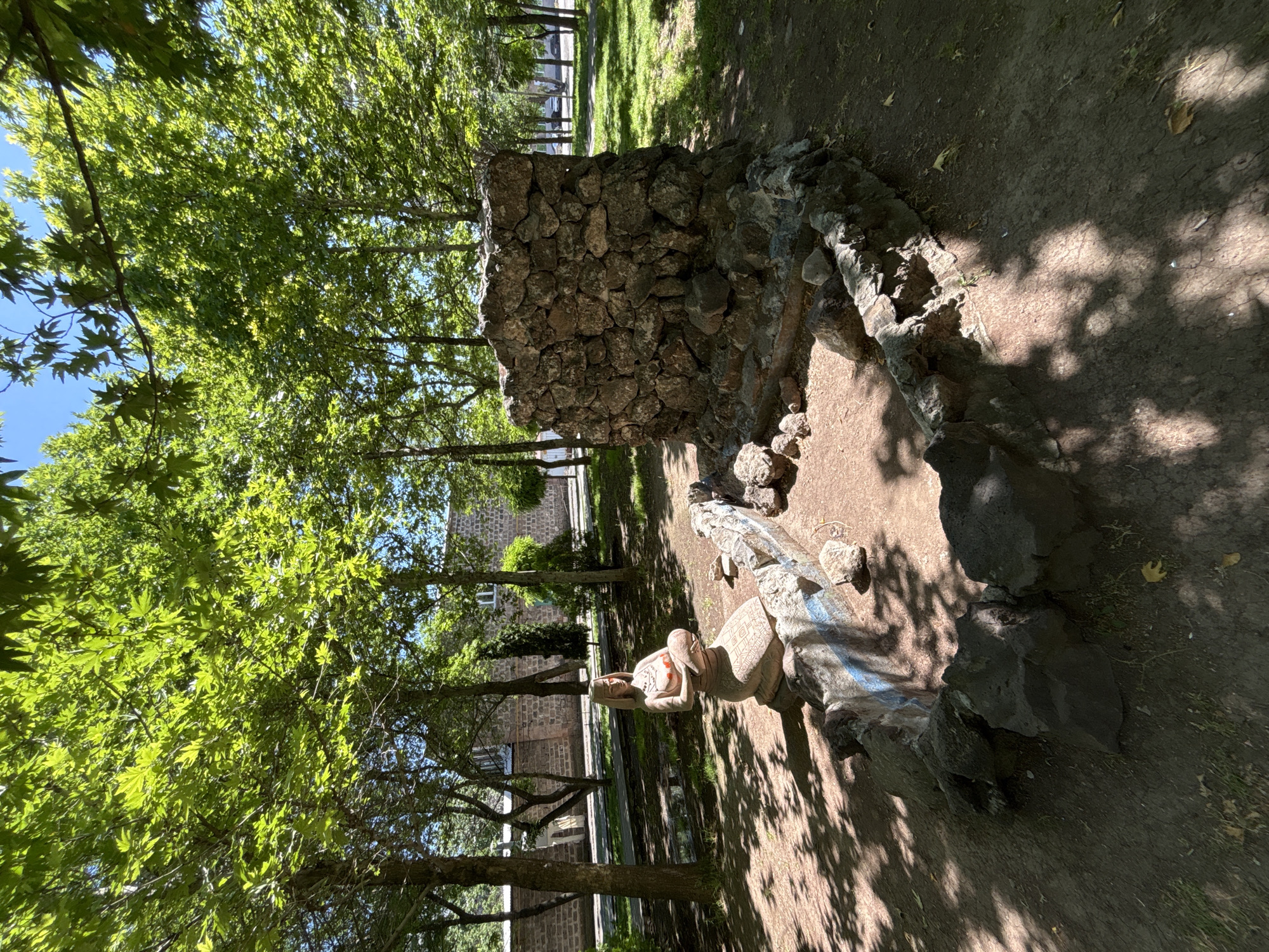 In Yerevan, Armenia, in a park setting with dappled sunlight filtering through lush green trees, a statue of a kneeling woman is situated next to a rough, stone-built structure. The statue, made of a light-colored material, depicts a woman in traditional dress, with her hands clasped around her chest. She has a headdress and painted red markings on her face and chest. Surrounding the statue and the stone structure is a low, irregularly shaped enclosure made of stones, with some of the stones painted with blue and white patterns. The ground is a mixture of dirt and some sparse vegetation. In the background, a stone building with arched windows can be seen, suggesting an urban park environment. The scene is peaceful and appears to be a monument or artistic installation within a public space.