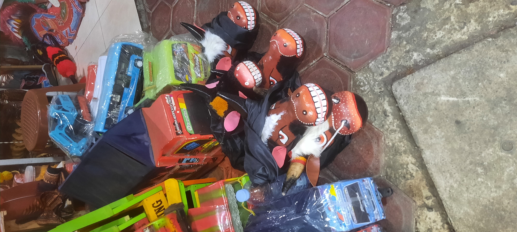 This image depicts a collection of traditional Indonesian "kuda lumping" horse masks and toy trucks displayed outdoors on a tiled surface, likely a street or market stall. The masks are elaborately decorated with painted faces, resembling horses or mythical creatures, and some are covered with black fabric. They are arranged in a group, with some appearing to be for sale. Several toy trucks, including a blue bus and a red truck, are also visible, some of them packaged in plastic. The environment suggests a bustling commercial area, possibly in Blitar, Indonesia, given the context of the kuda lumping tradition. The lighting and arrangement of objects indicate that this is a daytime scene. There are no people clearly visible in the frame.