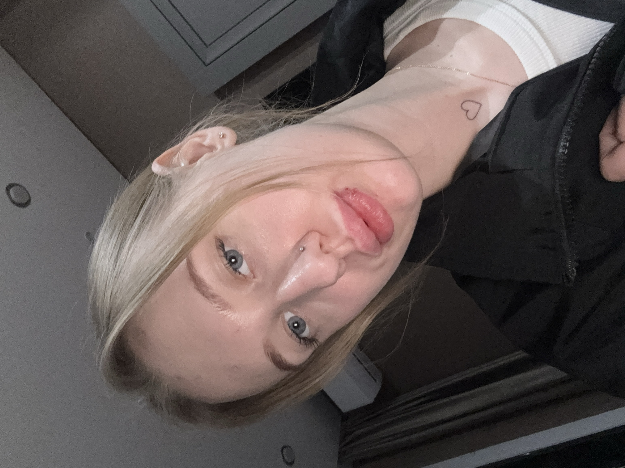 An image taken in the city of Azov, Russia, depicts a female individual in a selfie-style, close-up shot indoors. The subject, a woman with blonde hair and blue eyes, looks directly at the camera. She has a small silver stud piercing on her left nostril and her lips are intentionally pouted. A small, outline heart tattoo is visible on the left side of her lower neck. She is wearing a black zippered jacket over a white ribbed garment, and a thin chain necklace is visible. A small, light-colored stud earring is present in her right earlobe. The background shows an indoor environment with a white ceiling featuring a circular recessed light fixture, dark grey/brown walls, and a white wall-mounted air conditioning unit on the left. Dark vertical blinds or curtains are partially visible on the far left, and a dark panel or door frame is present on the right. The lighting suggests an artificial source from above.