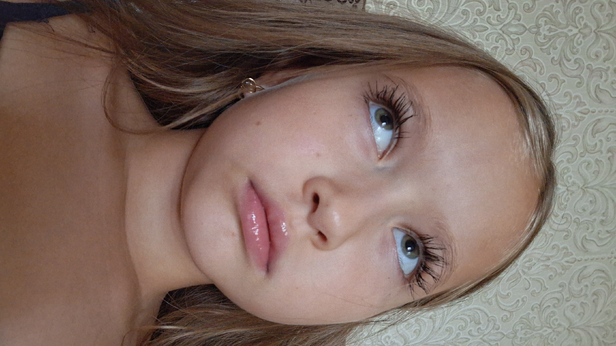 A young girl with light brown hair and green eyes looks up at the camera. She is lying down, with her head tilted to the side, and her hair is spread out around her. She is wearing a dark top and a small gold earring in her left ear. Her lips are slightly parted and appear to have lip gloss on them. She has long, dark eyelashes. The background is a patterned wallpaper in a light beige color. The lighting suggests it is daytime. The image appears to be a personal photograph, possibly taken indoors. The location is Semey, Kazakhstan.