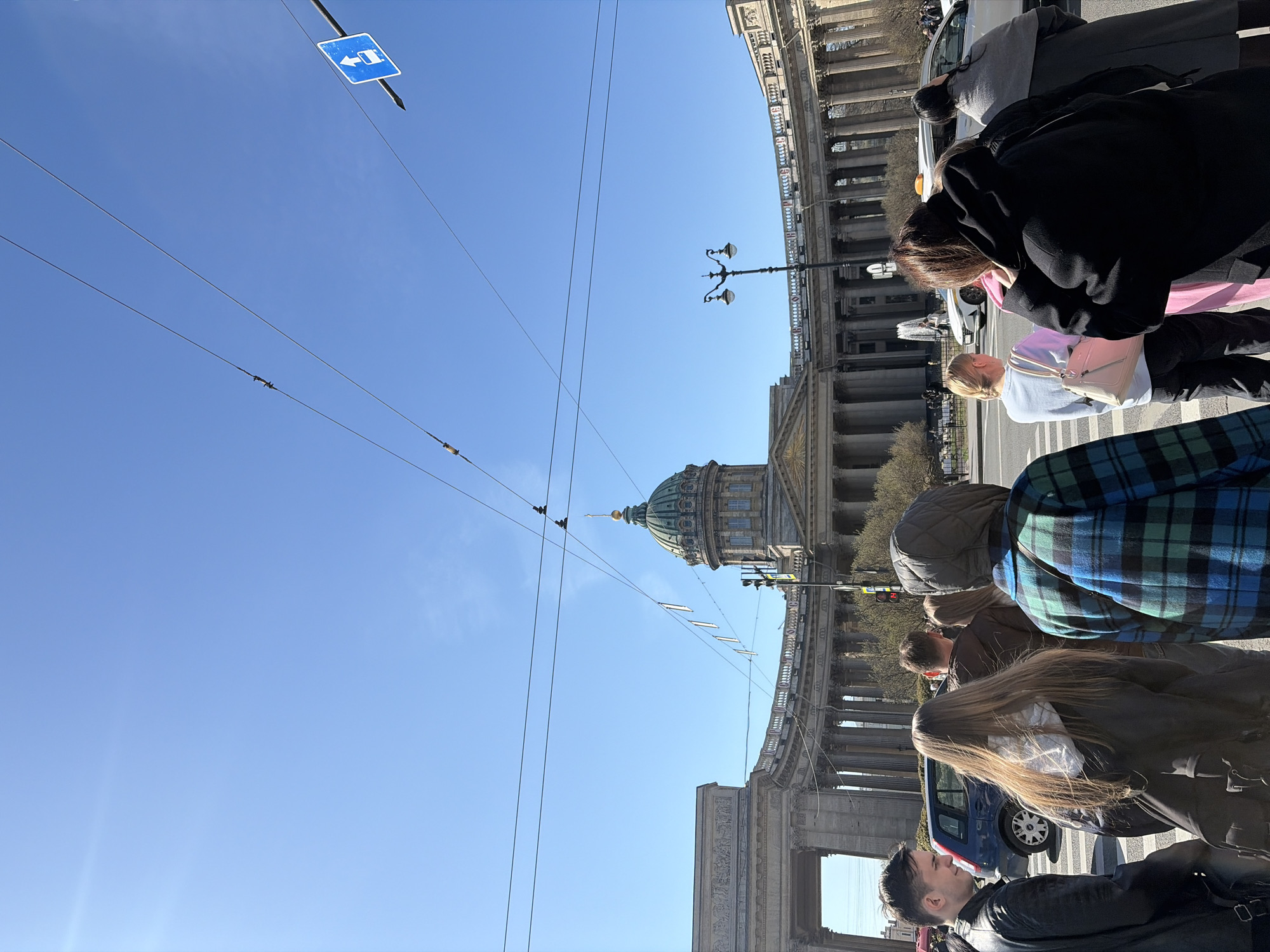 This daytime street scene captures a vibrant urban environment in Saint Petersburg, Russia, with the iconic Kazan Cathedral prominently featured. The image, originally rotated 90 degrees, depicts a clear, sunny day with a bright blue sky.
In the foreground, several pedestrians are visible, primarily from behind or the side, navigating a sidewalk alongside a street. On the left, a young man with dark hair wears a black leather jacket, looking towards the right. Next to him, a person with long, light brown hair and a dark jacket walks. Further back, a person in a distinctive blue and green plaid jacket with a black hood is seen. In the middle ground, a blonde woman in a light blue top and black pants carries a light-colored shoulder bag, while behind her, another person in a black coat and pink top looks downwards.
The background is dominated by the majestic neoclassical architecture of the Kazan Cathedral, easily identifiable by its grand colonnade and the large, green-domed cupola. Overhead, numerous thin wires, likely for trolleybuses or power, crisscross the sky. An ornate black streetlamp with multiple lights is visible on the right side, positioned against the cathedral. A blue square road sign in the upper left features a white arrow pointing left and a white symbol resembling a car or public transport stop. Scattered parts of cars, including a white sedan and a blue car, are visible on the street, indicating ongoing traffic. The scene conveys a typical, bustling day in the city, with no specific event unfolding, just the everyday flow of life.