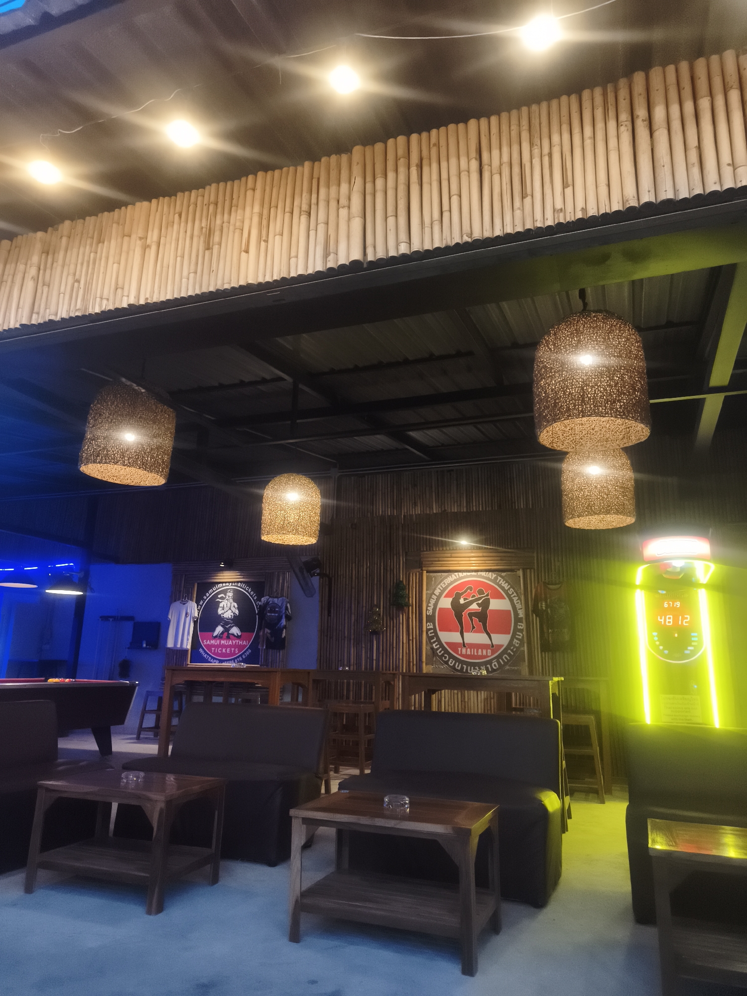 The image shows the interior of a bar or entertainment venue in Baan Chaweng, Thailand. The ceiling has bamboo accents, with woven pendant lights hanging. Furnishings include dark-colored sofas, wooden tables, a pool table, and a brightly lit, possibly coin-operated, game or entertainment device. Wall decor consists of promotional posters or signage for "Samui Muay Thai Tickets" and "Samui International Muay Thai Stadium". The setting appears casual and inviting.