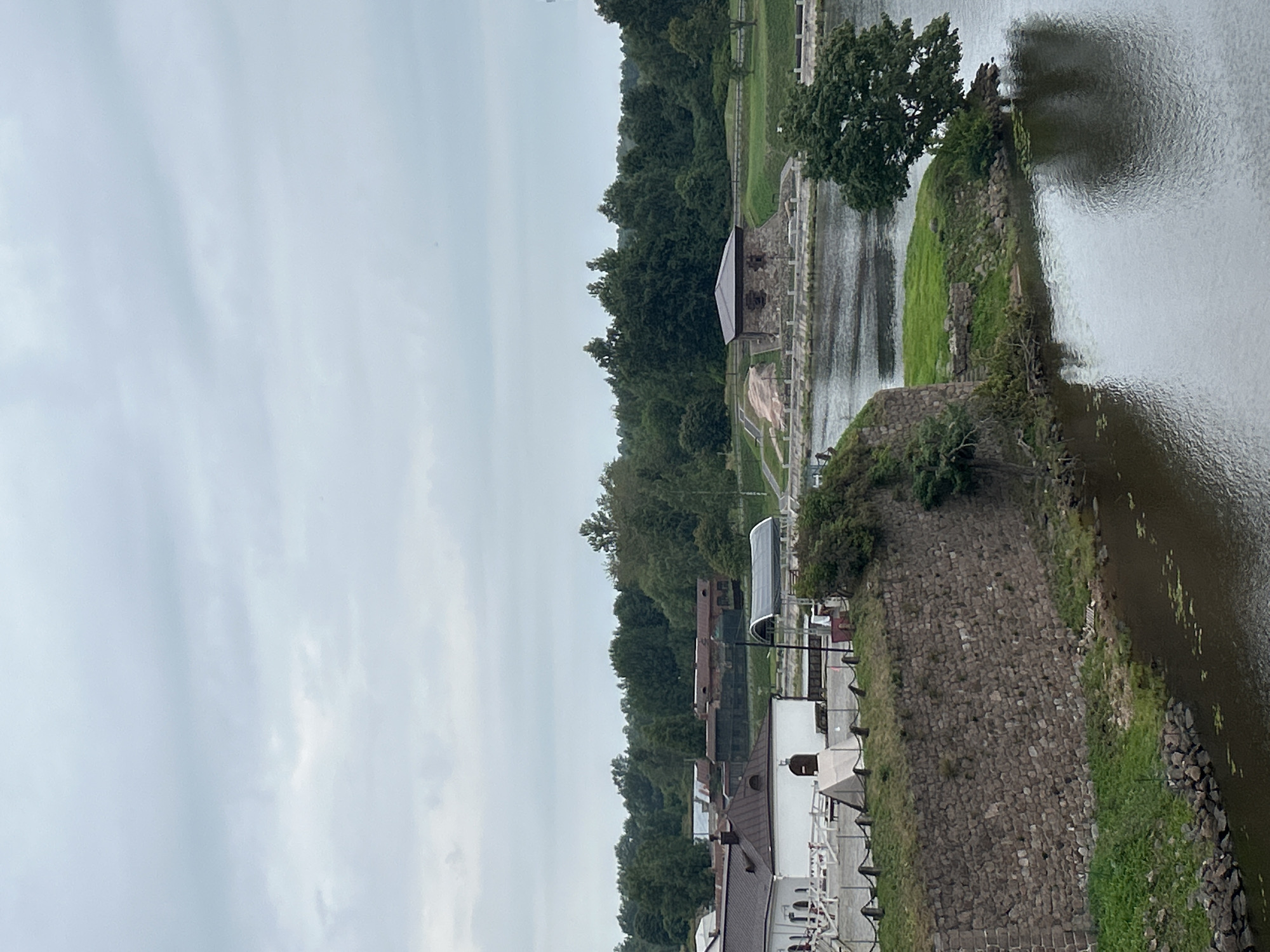 The image captures a serene waterside scene within Vyborg, Russia, featuring the Vyborg Castle's historic stone ramparts and fortifications. In the foreground, a rough-hewn stone wall, covered in patches of green grass, forms the edge of the fortress. A body of dark water, possibly a moat or bay, reflects the overcast sky and surrounding greenery.
Beyond the wall, a white building with a dark-tiled roof is visible, part of the castle complex. Further back, a cluster of older buildings with corrugated metal roofs suggests historical structures within the city. Across the water, a quaint, stone-built structure with a dark, pitched roof stands on a grassy knoll, appearing to be a small tower or auxiliary building.
The background is dominated by a dense forest of lush green trees, characteristic of the Russian landscape. The sky is a uniform pale gray, indicating an overcast day with no visible sun. The overall atmosphere is one of quiet historical grandeur, with the natural environment playing a significant role in the composition. There are no people readily identifiable in the image. The scene is static, devoid of any apparent action or interaction.