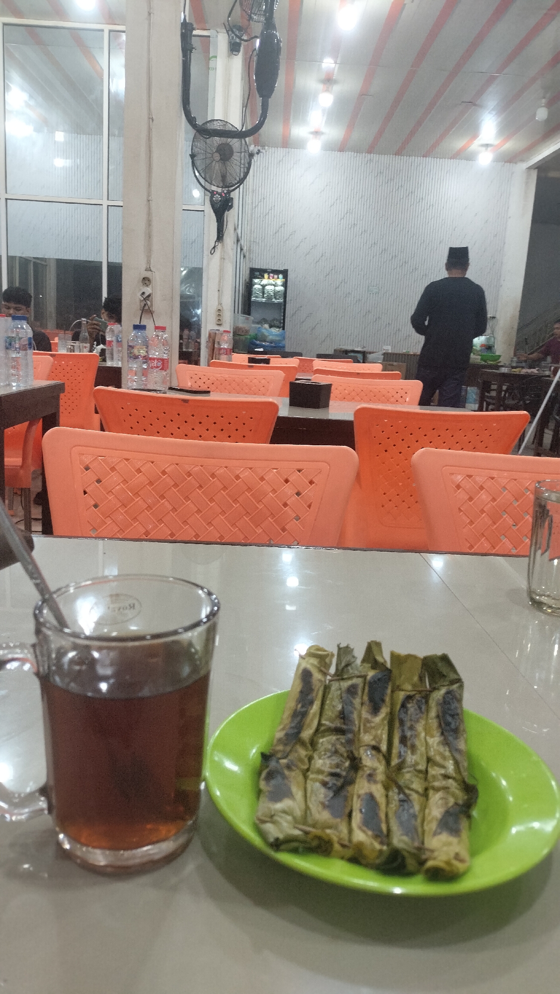 The image depicts a dining area, possibly a restaurant, in Sigli, Indonesia. In the foreground, on a table, is a glass mug filled with a dark liquid, likely tea, and a green plate containing several grilled food items wrapped in leaves. The background reveals a spacious interior with numerous empty orange plastic chairs arranged around tables. A ceiling fan is visible, along with strip lighting on the ceiling. Further back, a person wearing a black shirt and a peci hat is standing and facing away from the camera. The environment appears to be indoors, with large windows or glass panels on the left side. The overall atmosphere suggests a casual dining setting.