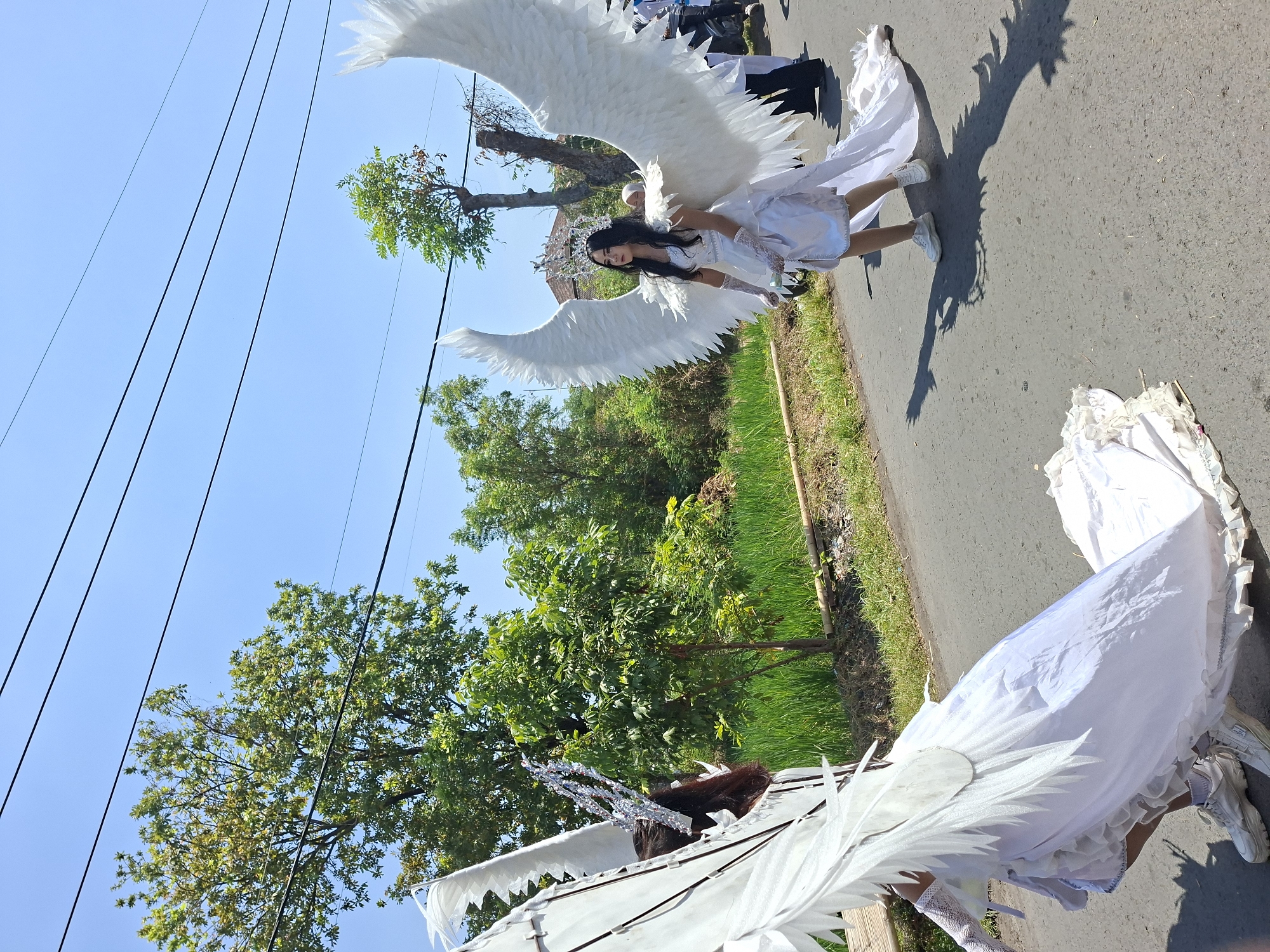 Two individuals are walking on a paved road in Tongaswetan, Indonesia, dressed in elaborate white costumes resembling angels. The person on the right has large, white feathered wings extending from their back and is wearing a white dress, gloves, and a crown. The person on the left also has large white feathered wings and a crown, with their back mostly visible. Both are wearing white sneakers. They are walking on a road with a grassy area and trees on the left, and more trees and structures in the background. The sky is clear and blue. The scene appears to be outdoors during the day, likely part of a parade or event.