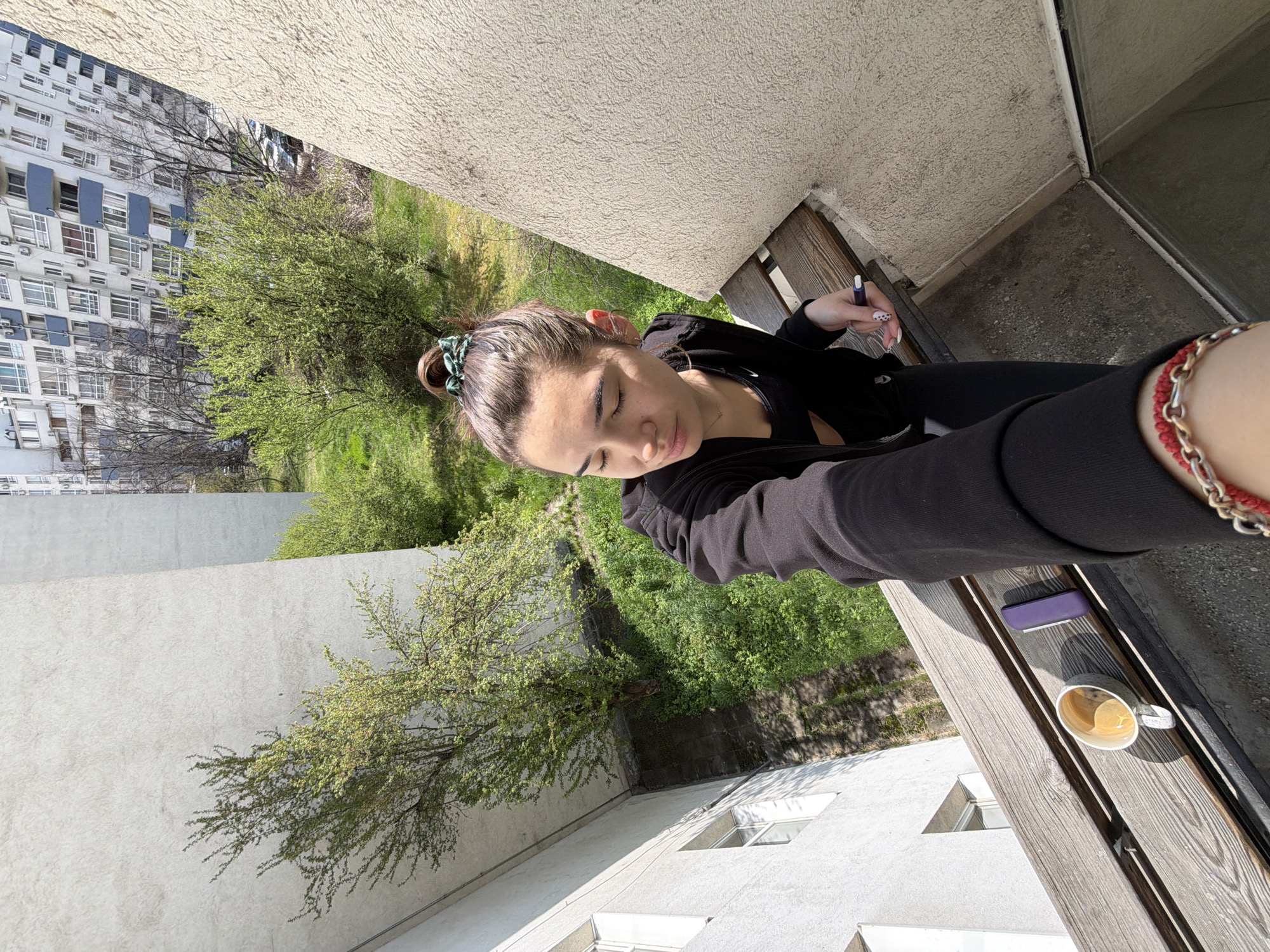 A medium, eye-level shot depicts a woman in a black hooded sweatshirt seated on a wooden balcony. She has dark hair tied in a bun with a green scrunchie. She holds a pen in her right hand. On the balcony next to her are a purple case and a white coffee cup with coffee inside. The woman's left arm is outstretched towards the viewer, with a red and gold bracelet on her wrist.
The background reveals a cityscape, with lush green trees and a high-rise apartment building with white walls and blue-grey accents on the left. The architecture suggests this location is potentially in Sofia, Bulgaria, given the typical style of residential buildings in that area. The angle and layout suggest the image was taken from a high vantage point. The light indicates a sunny day.