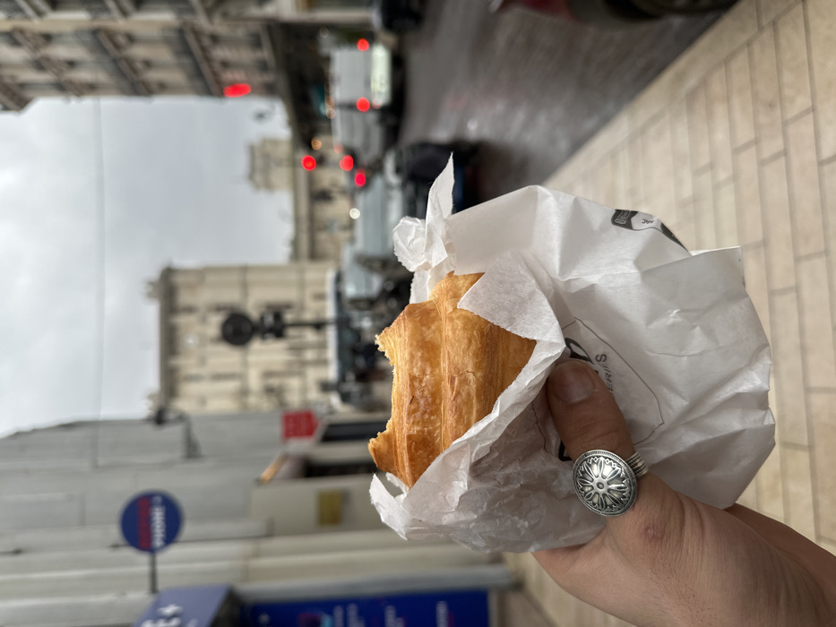 The image depicts a first-person perspective shot on a street in Vincennes, France, under an overcast sky. In the foreground, a left hand holds a partially consumed golden-brown croissant, wrapped in crinkled white paper, from which a bite has been taken from the left side. The hand wears a large silver ring on the ring finger, featuring a circular, detailed relief pattern. Partially visible text on the white paper suggests "BOULANGERIE".
The background is out of focus, showing an urban street scene. The asphalt road surface appears wet, indicating recent precipitation. Various vehicles, including passenger cars and a white utility van, are present on the road, with several red tail lights illuminated. To the left, a building facade displays a blue sign with white text, partially visible as "PHONE+". An elevated train track structure, characteristic of an RER line, spans across the mid-ground, with an indistinct train visible beneath it. Beyond the street, multi-story buildings with light-colored stone facades and classic Parisian architectural elements line the view, including a prominent, large building with numerous windows in the distant background. A black traffic light pole is visible in the mid-ground.