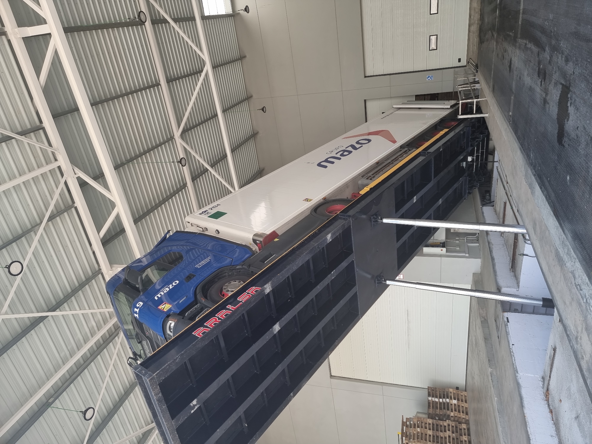 An eye-level, medium shot captures a blue truck and its trailer on a tilted loading dock ramp inside a warehouse in Alaquàs, Spain. The ramp, made of black metal, angles upward towards the left of the frame. The truck's cab is visible on the left side of the ramp, displaying the number "119" and "GRUPO MAZO" branding. The trailer, white with "GRUPO MAZO" logo in large blue letters, extends to the right. The warehouse interior features a white corrugated metal ceiling with exposed beams and industrial lighting fixtures. To the left of the ramp, stacks of wooden pallets rest on the concrete floor. The background shows a large, closed white roller door. The lighting is bright, suggesting an indoor, possibly industrial or commercial, setting.