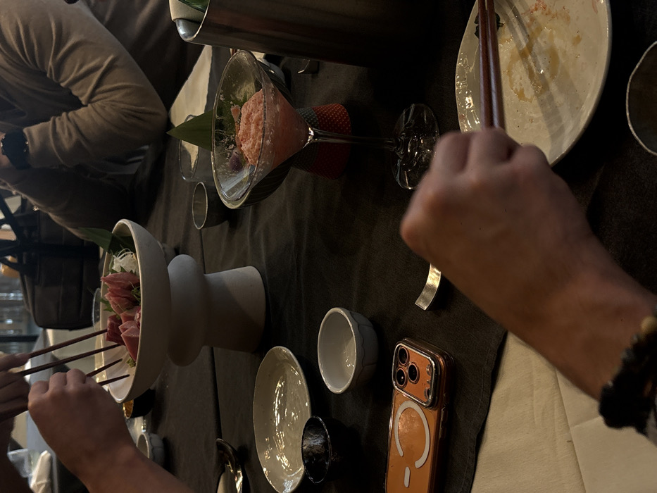A dining table is set with various dishes and utensils. A person is holding chopsticks near a white bowl filled with food. Another person is visible, and there is a martini glass filled with a pink mixture. An iPhone in an orange case sits on the table beside a small bowl. An empty plate with chopsticks rests near a silver spoon. A metallic container is in the background.