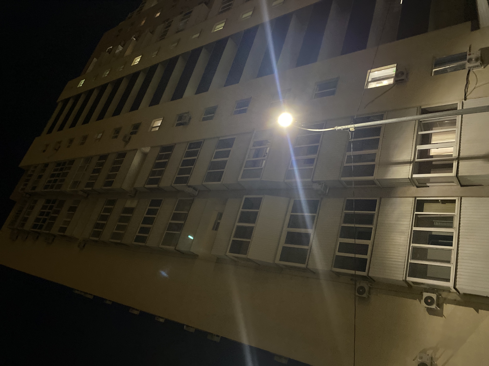 The media file is a low-angle, wide shot of a modern, multi-story building at night. The building's facade is a light color, likely beige or cream, and features numerous windows of varying sizes and configurations. Some windows are illuminated from within, suggesting occupancy. Vertical, dark architectural elements punctuate the facade, creating a striped effect. A bright light fixture, possibly a street lamp, is visible in the upper middle portion of the image, casting a strong glare and lens flare across the scene. Air conditioning units are mounted on the exterior walls beneath some of the windows. The ground below is not visible, and the surrounding environment is dark, indicating it's nighttime. The overall impression is one of an urban or semi-urban setting, with a focus on the architecture of the building. There is no visible text in the image.