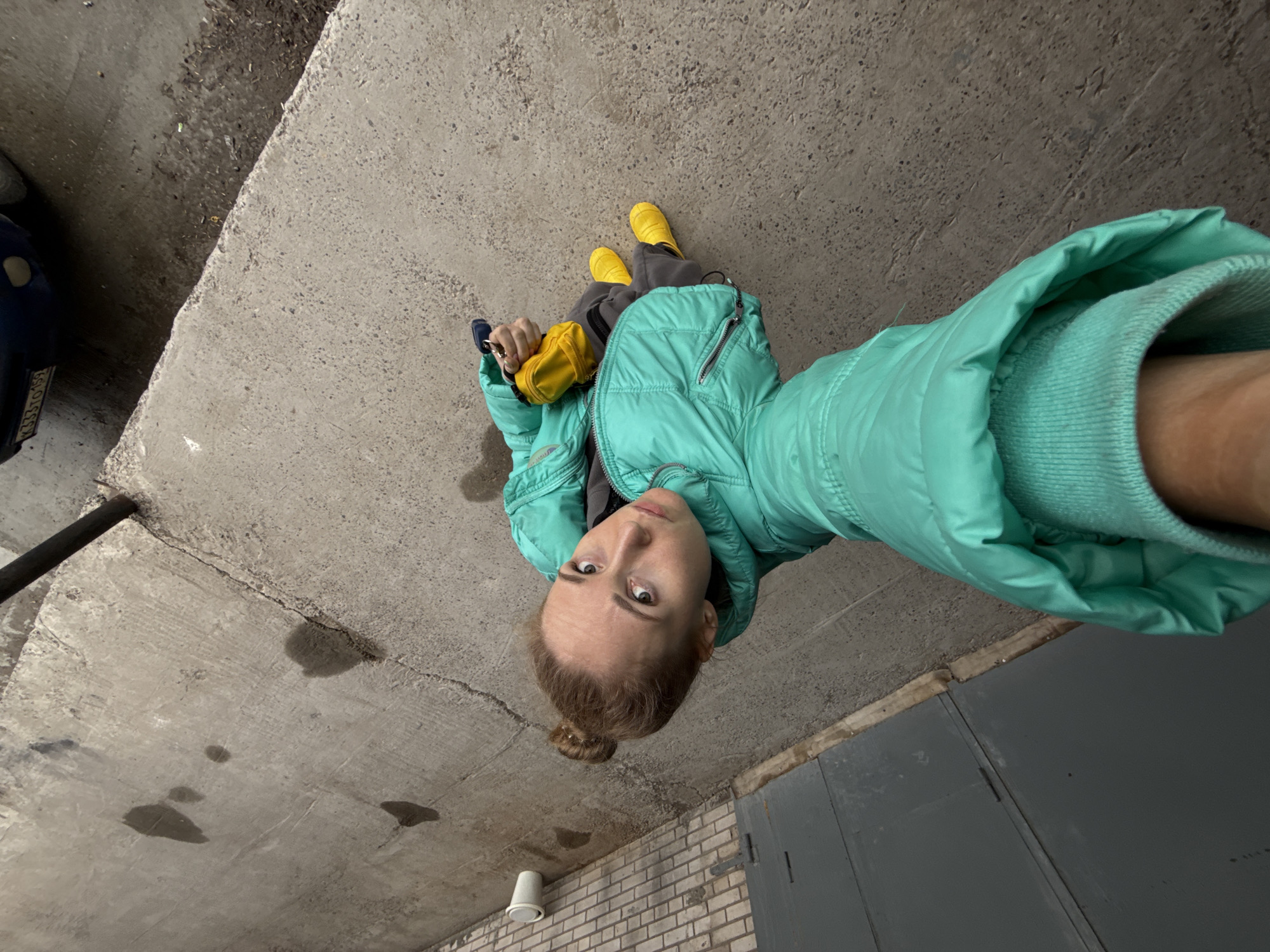A low-angle photograph captures a person, likely a woman, looking directly into the camera from an overhead perspective. She is standing on a rough concrete surface, dressed in a vibrant teal puffer jacket, grey sweatpants, and distinctive bright yellow rain boots. In her hand, she holds a small yellow pouch with keys attached.
The environment suggests an urban or industrial setting, possibly a covered outdoor area, an underpass, or a multi-story parking garage in Chaikovsky, Russia. The "ceiling" above her is also made of concrete, showing cracks and various dark, wet stains, indicating damp conditions or water leakage. More wet patches are visible on the ground where she stands.
To the left of the frame, a partial view of a blue car's front fender and headlight is visible, along with a license plate displaying "K333TO 159". The "159" is a Russian region code for Perm Krai, where Chaikovsky is located. To the right, a dark grey garage door can be seen, and a brick wall with a discarded white plastic cup is situated closer to the bottom right.
The person's expression is neutral as she poses for the camera. The wet ground and rain boots suggest it might be or has recently been raining, while her puffer jacket indicates cool weather. The lighting suggests it is daytime.