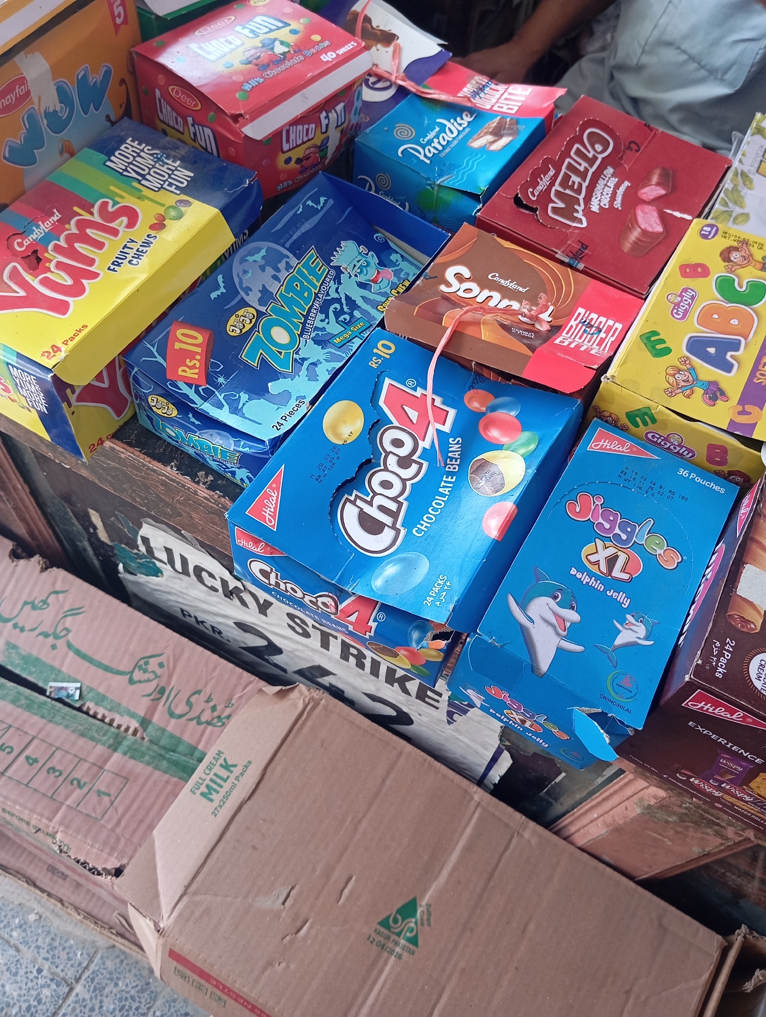 The image shows a close-up, high-angle view of a variety of colorful confectionery boxes and packaging, possibly from a shop in Mansehra, Pakistan. The items are stacked and arranged haphazardly, suggesting a retail environment. Some visible brands include "Candyland Yums," "Zombie," "Choco4," "Mello," "Sonnet," and "Jiggles XL." There are also boxes for "FULL CREAM MILK." A torn cardboard box with the text "LUCKY STRIKE" is prominent in the lower portion of the frame. The overall impression is one of a busy, somewhat disorganized display of sweets and dairy products.