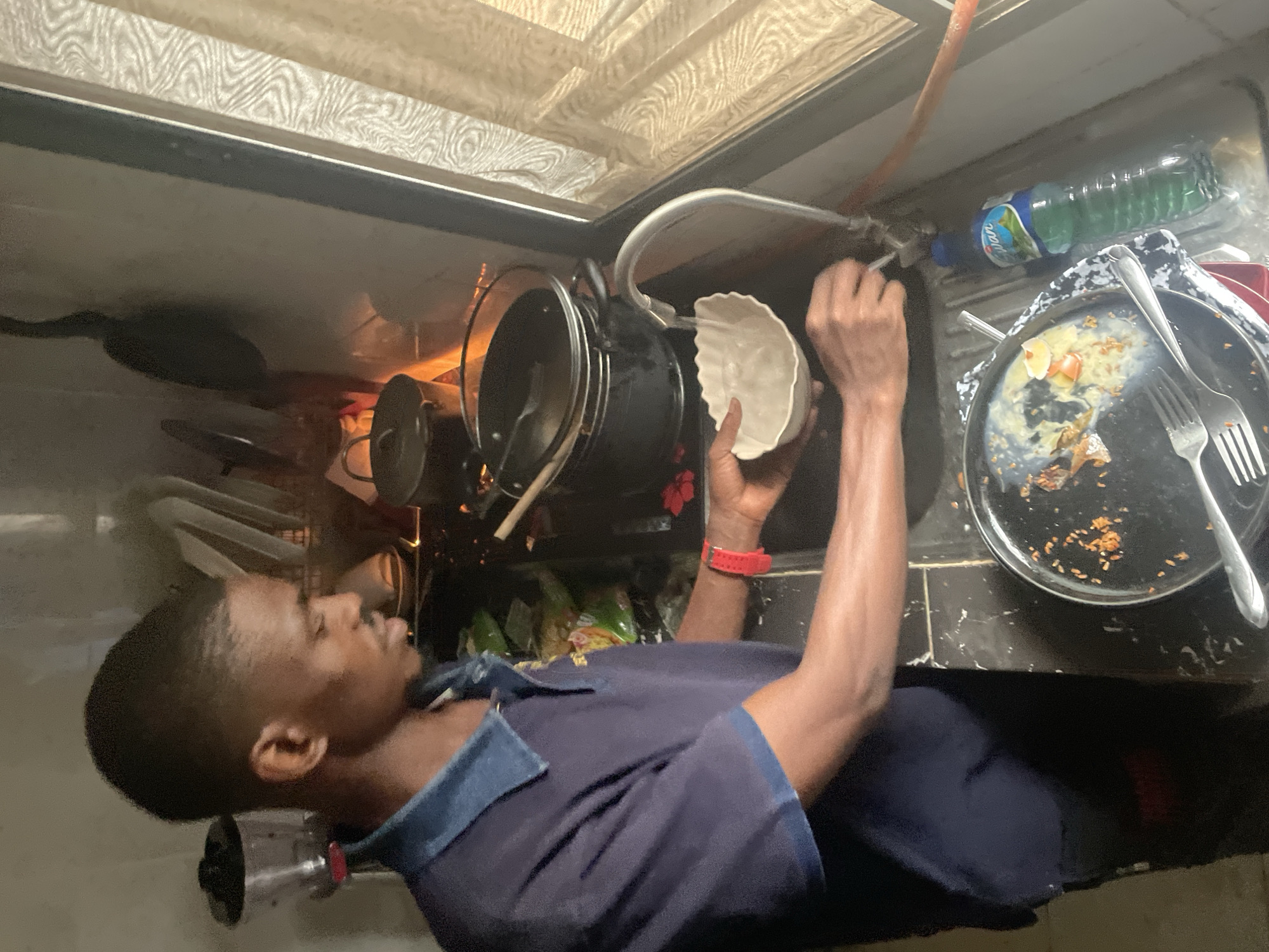 A man stands at a sink washing dishes. He is holding a white, scalloped bowl under the running water from the faucet. To his right is a dirty plate with leftover food and utensils. Behind the sink, a bottle of green liquid dish soap is visible. There are pots and other dishes in the sink. The man has dark hair and is wearing a dark blue collared shirt. A red wristband is visible on his left wrist. The location is likely in Abuja, Nigeria, based on the context of the question.