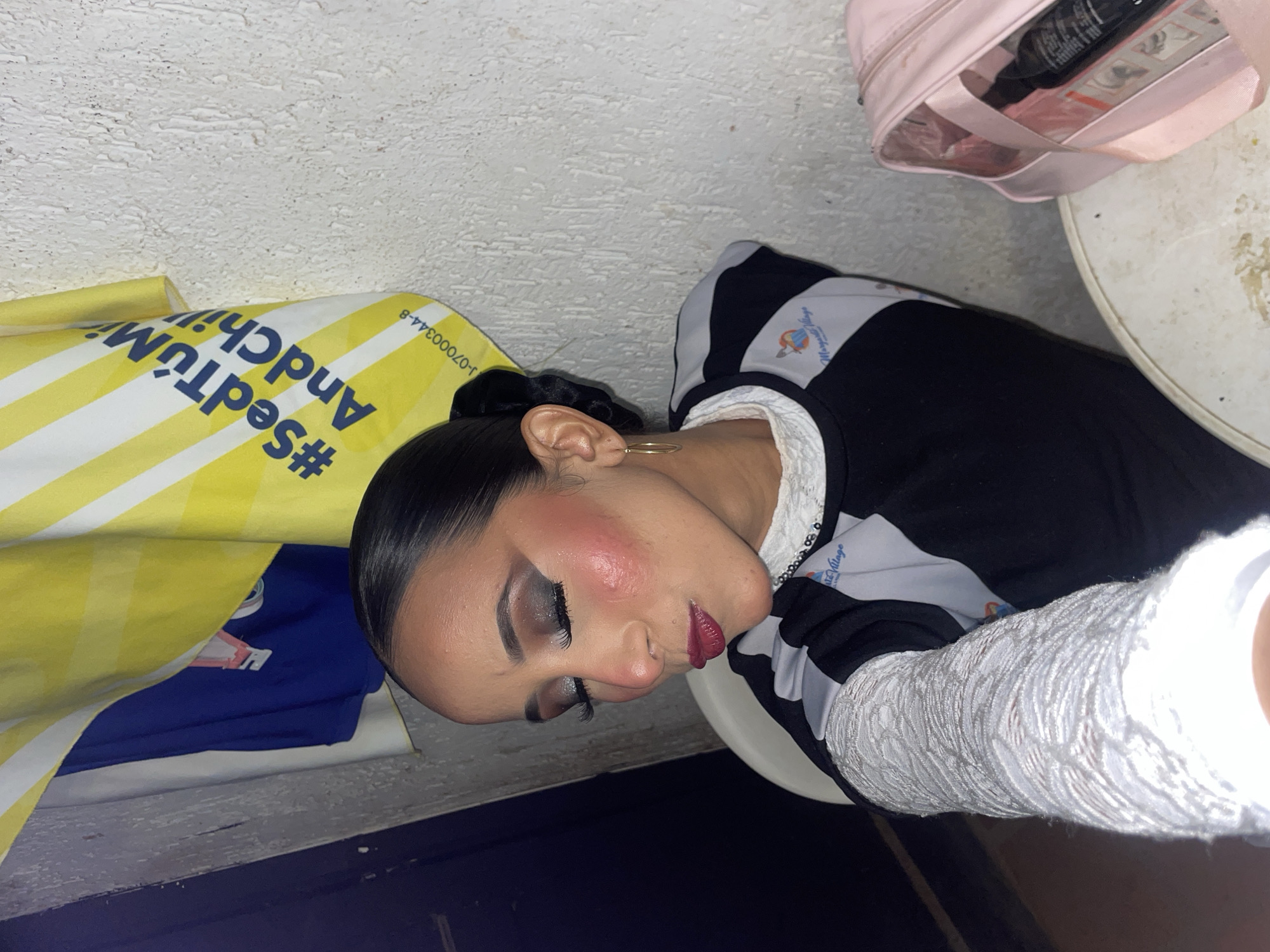 The image is a medium shot of a young woman with dark hair pulled back, wearing elaborate makeup including heavy eyeshadow, false eyelashes, and dark lipstick. She has prominent blush on her cheeks and is wearing a black and gray top with white lace accents and a gold hoop earring. She is seated in front of a textured white wall, with a blue shirt and a yellow and white striped banner or towel behind her. A pink bag is visible to the right.
This scene appears to be inside a room, possibly a dressing room or a backstage area, given the makeup and attire. The woman's focused expression suggests she is either posing for a photo or preparing for an event. The overall setting is somewhat cluttered, with the background elements suggesting a less formal environment.
Given the specific request to include the location in Porlamar, Venezuela, and the details visible in the image, it is not possible to definitively confirm this location based solely on the visual information provided. There are no explicit markers such as street signs, landmarks, or local signage that would confirm Porlamar, Venezuela as the setting.