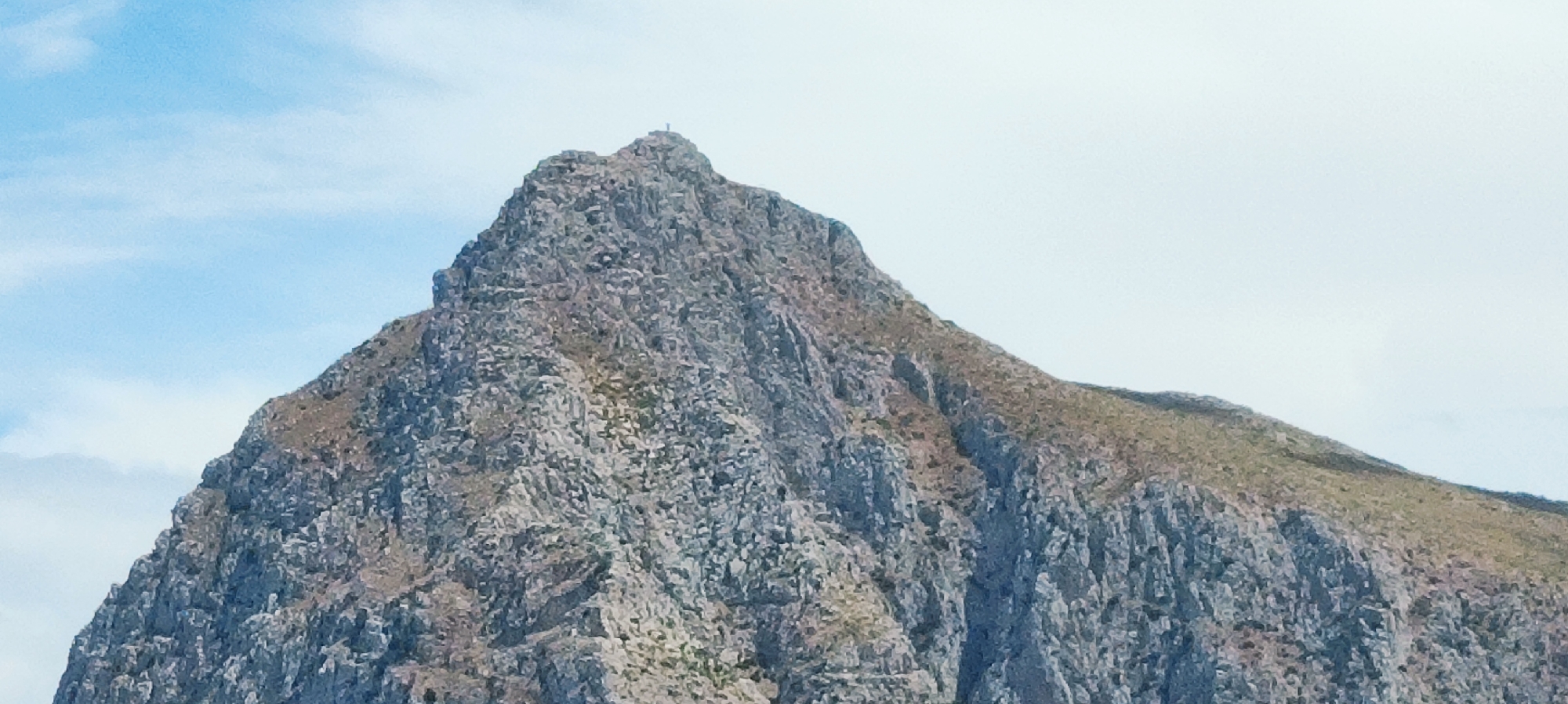 A close-up, low-angle shot showcases the rugged, rocky summit of Monte Monaco in San Vito Lo Capo, Italy. The mountain dominates the frame, its steep, jagged face covered in grey and brown rocks with sparse patches of dry vegetation. At the very peak, a small, solitary figure stands, possibly a hiker or a cross, silhouetted against the bright, hazy sky. The sky above is a pale blue, with faint white clouds. The image captures the imposing natural beauty of the mountain and its summit.