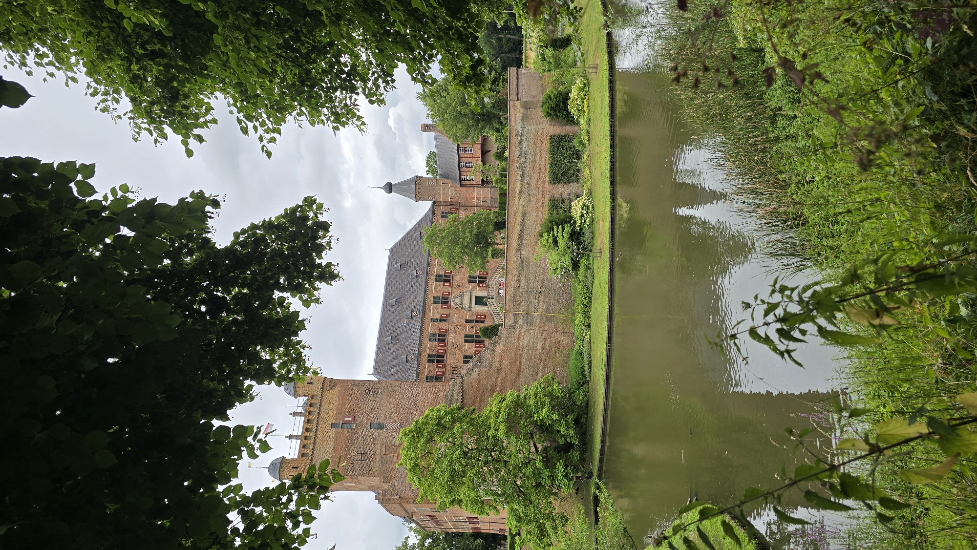 The image shows a castle, likely Huis Bergh in 's-Heerenberg, Netherlands. The castle is constructed of brick and features several towers and a main building with a pitched roof. Red shutters are visible on many of the windows. The castle is surrounded by a moat filled with murky green water, and a brick wall encloses the castle grounds. Lush green trees frame the top and sides of the image, with foliage casting shadows. In the foreground, there is a pond or moat with reflections of the castle and surrounding trees. The sky is overcast.