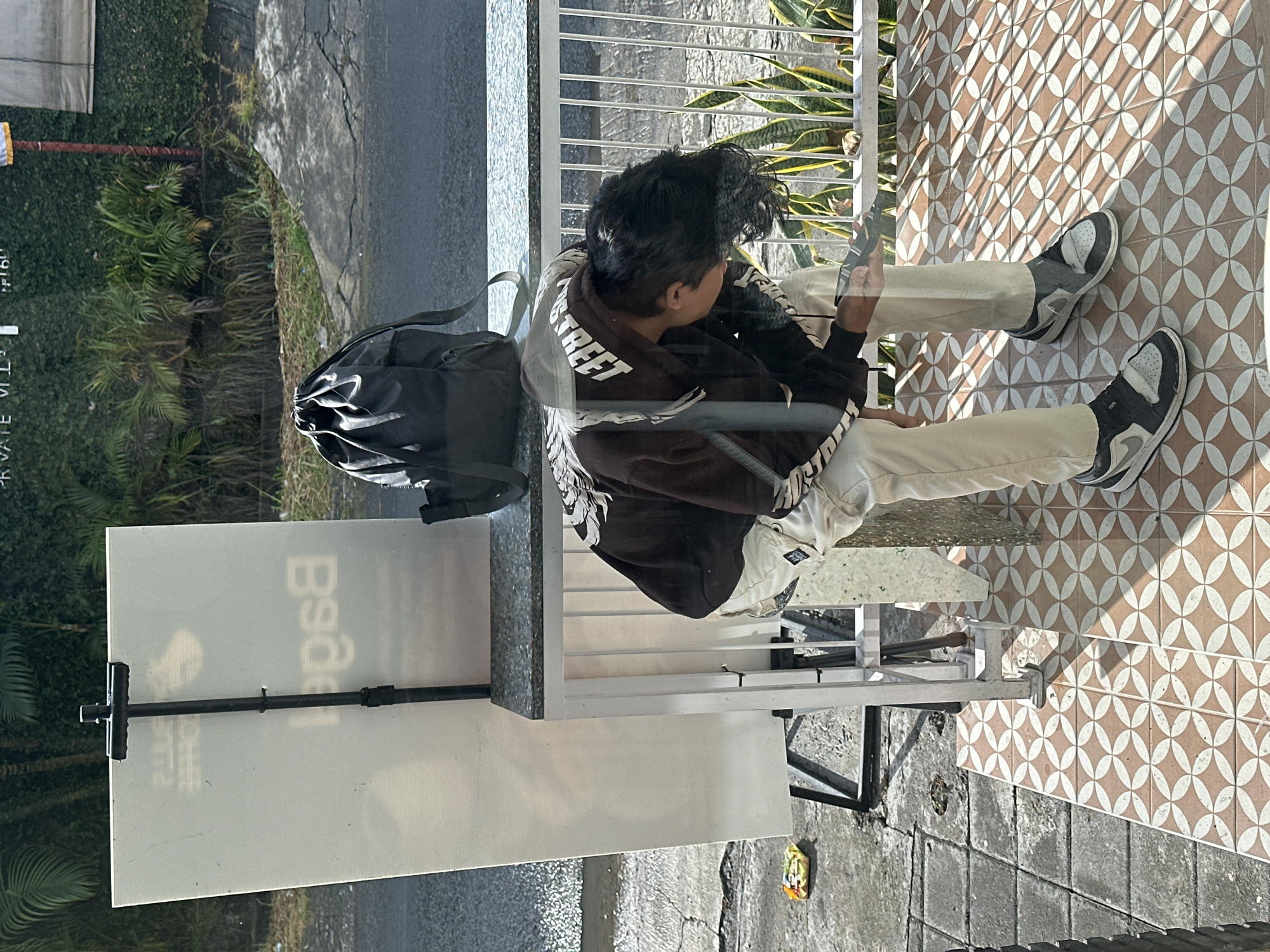 A young male is bent over, looking down at his phone, in front of a white board with text, possibly a menu or advertisement, at an outdoor establishment. He is wearing a dark brown hoodie with white lettering, light-colored pants, and black and white sneakers. A black backpack rests on a metal railing behind him. The ground is paved with geometric tiles and concrete. Lush green foliage is visible in the background, suggesting a tropical environment. The scene appears to be a street-side cafe or shop in North Kuta, Indonesia.