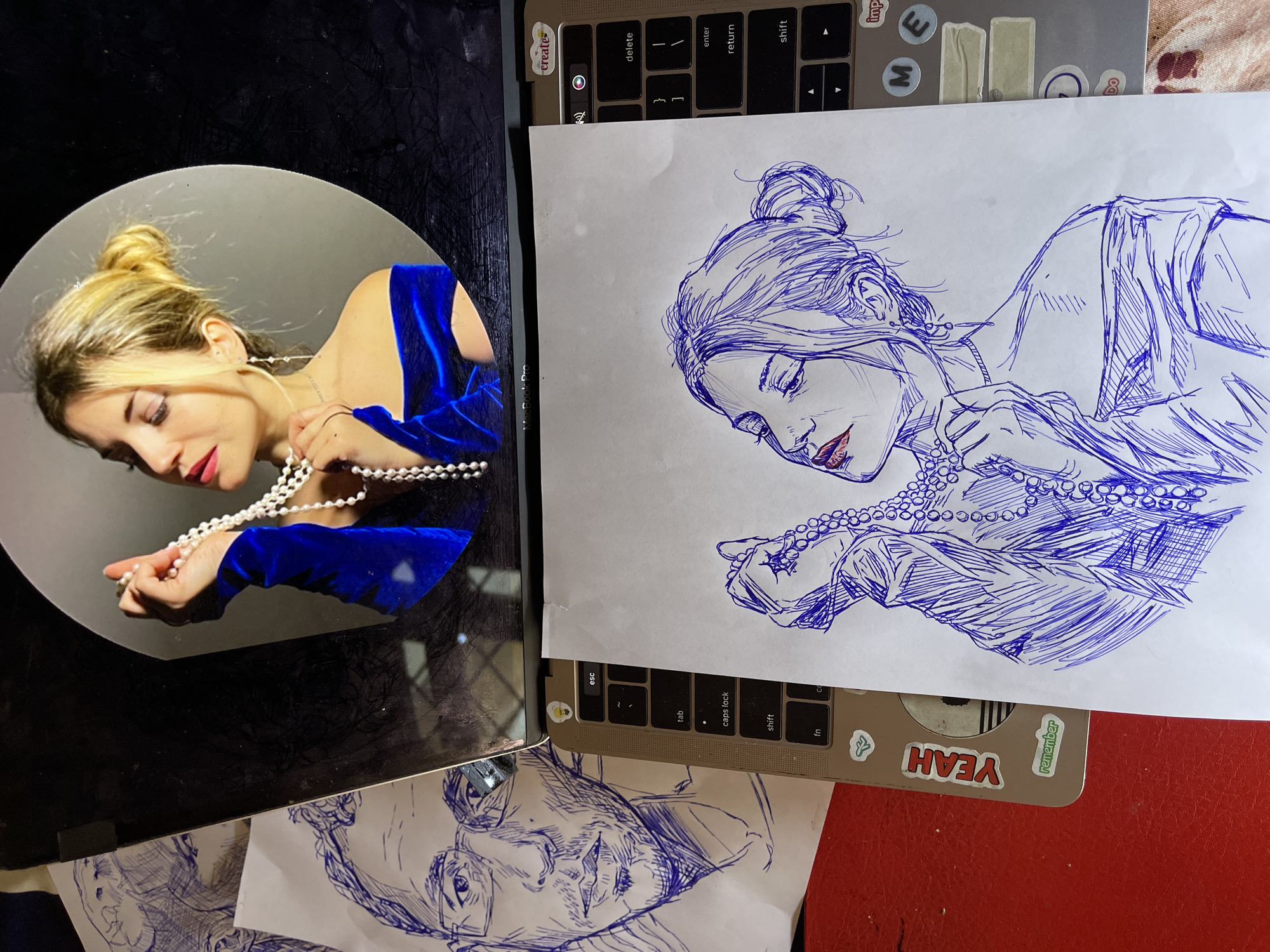 The image shows a close-up of a laptop with a drawing on a piece of paper in front of it. The drawing is a sketch of a woman wearing a pearl necklace and a blue off-the-shoulder top. The woman in the drawing has her hair in a bun. To the left of the drawing is another sketch of a man's face. The laptop screen reflects an image of a woman also wearing a pearl necklace and a blue off-the-shoulder top, which appears to be the subject of the sketches. The laptop is on a red surface, possibly a desk or table. The overall scene is a workspace with art supplies. There is no information in the image that suggests the location is the city of Jos, Nigeria.