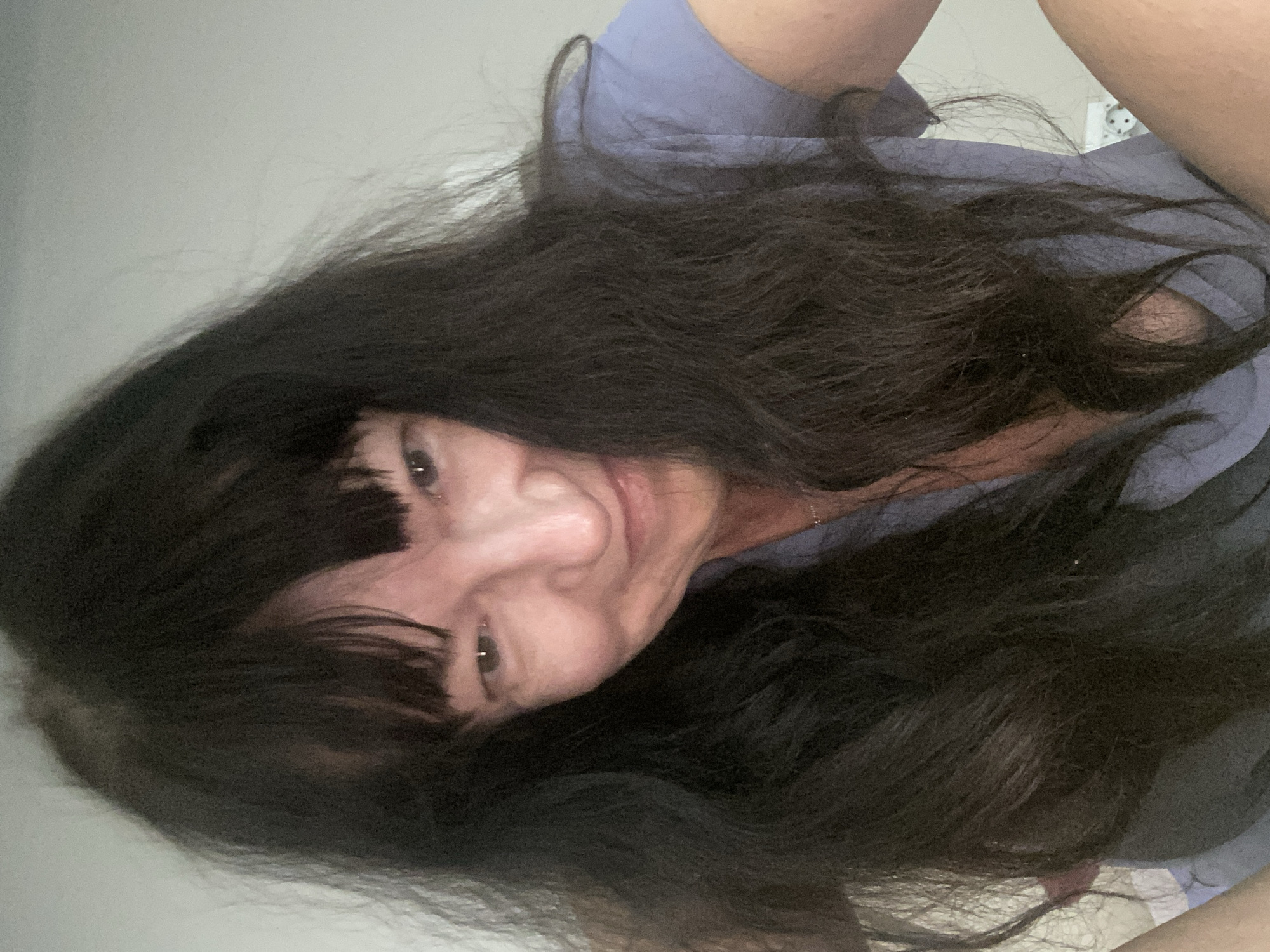 A selfie of a woman with long, dark, wavy hair and bangs. She is wearing a light blue t-shirt and looking at the camera with a slight smile. The background is plain and light-colored, suggesting an indoor setting. The image does not contain information about the city of Novi Petrivtsi, Ukraine.
