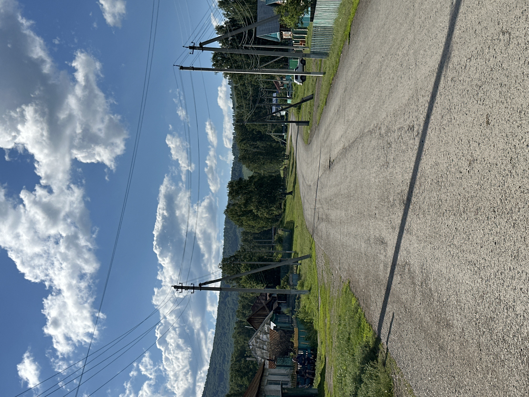 A gravel road curves through a rural village under a partly cloudy blue sky. On the left, houses with green fences and wooden structures are visible, with two people sitting on a bench in front of one house. Telephone poles with wires line the road. Trees and a forested hill form the background. The road is somewhat damaged with some potholes.