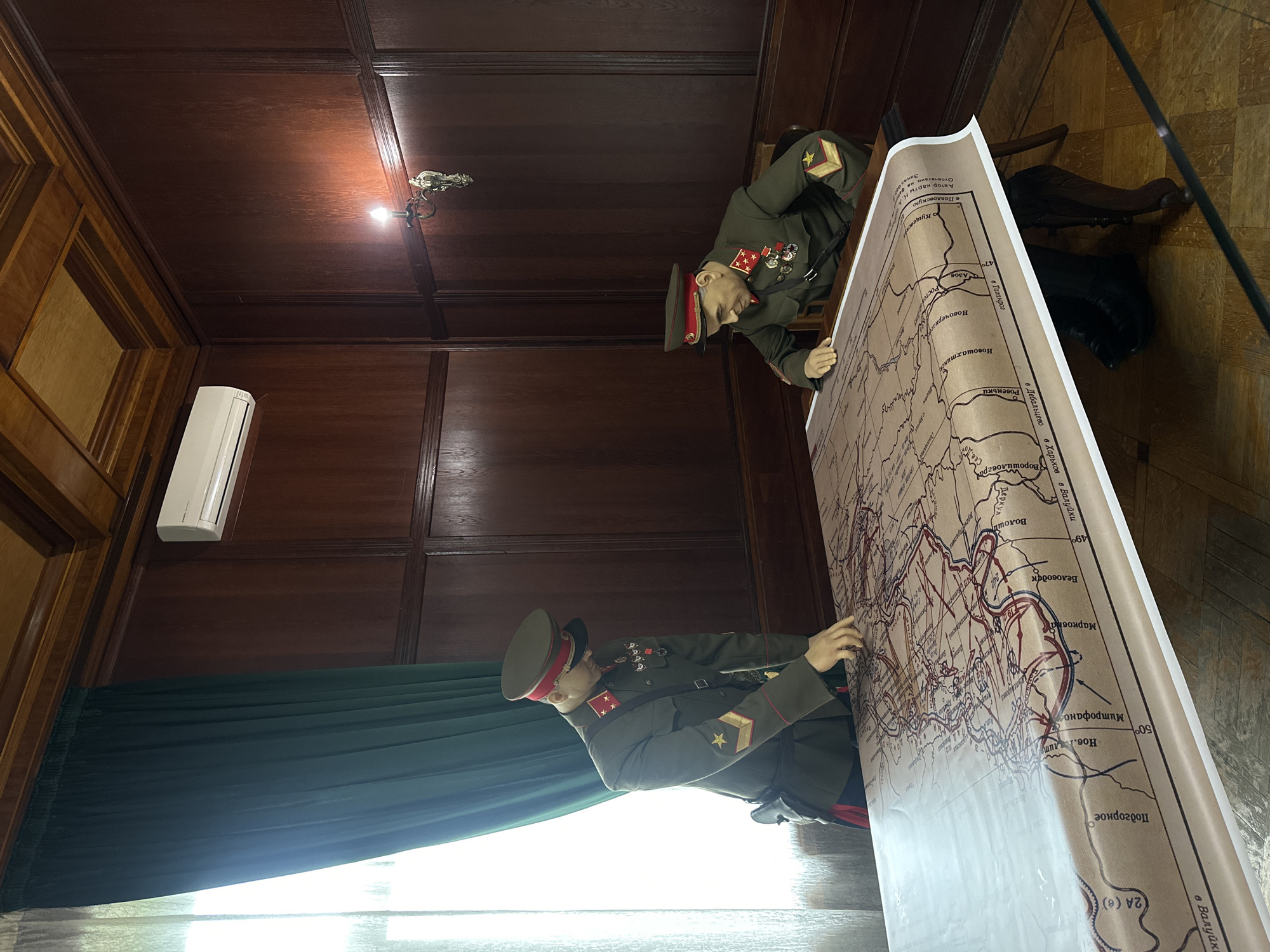 The image captures a scene within a room adorned with wood-paneled walls and a hardwood floor. A dark green curtain drapes near a window, and an air conditioning unit is mounted on the wall. The focal point involves two mannequins dressed in Soviet military uniforms, positioned on either side of a large map. The mannequins appear to be examining the map, which details geographical locations such as "Волоши," "Беловодск," and "Марковна." The text on the map includes phrases like "Автор карты Н.А" and "Отпечатано." The location is likely a museum or historical exhibit in Sochi, Russia, given the subject matter and the style of display.