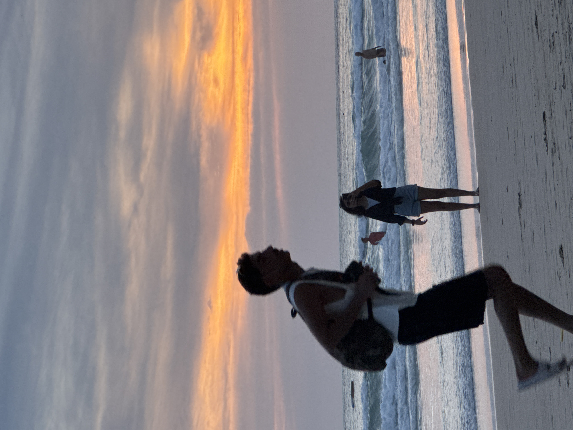 An outdoor, eye-level, medium-shot photograph captures a beach scene in Karon, Thailand, during a vibrant sunset. The sky is filled with bands of orange and pink clouds, transitioning to muted blues and grays higher up. The ocean is active with moderate waves crashing onto the shore, leaving a foamy surf line.
In the foreground, a young man is captured mid-stride, running from left to right across the sandy beach. He is silhouetted against the sunset, wearing a white tank top and dark shorts, with a backpack slung over his left shoulder. His focus appears to be forward, and he is holding a camera in his right hand.
Further back, near the water's edge, a young woman stands facing the camera. She is wearing a dark jacket over a light-colored top and denim shorts. She is holding a smartphone in her hands, seemingly taking a photograph or recording.
In the ocean, a person is standing waist-deep in the water, facing away from the camera. Another person is visible in the distance, further out in the sea, near a small object that could be a buoy or a small boat. The beach itself is sandy, with some subtle ripples and shadows indicating the uneven terrain. The overall mood is tranquil and evocative of a late afternoon or early evening on a tropical beach.