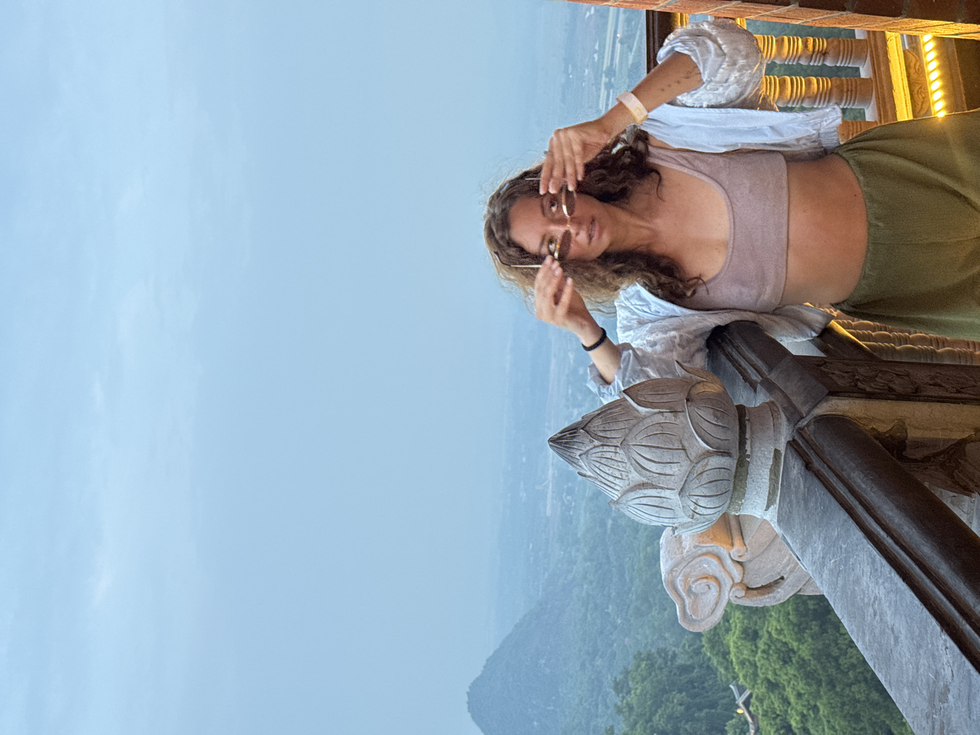 In Tây Hoa Lư Ward, Vietnam, a woman with curly brown hair stands on a balcony overlooking a misty valley. She is wearing a light pink crop top, an open light blue shirt, and olive green pants. She is holding a pair of sunglasses up to her eyes. The balcony railing is made of stone and features intricate carvings, including a lotus flower design. In the background, a mountain peak emerges from the fog, and lush green trees cover the slopes. The sky is overcast and a pale blue.