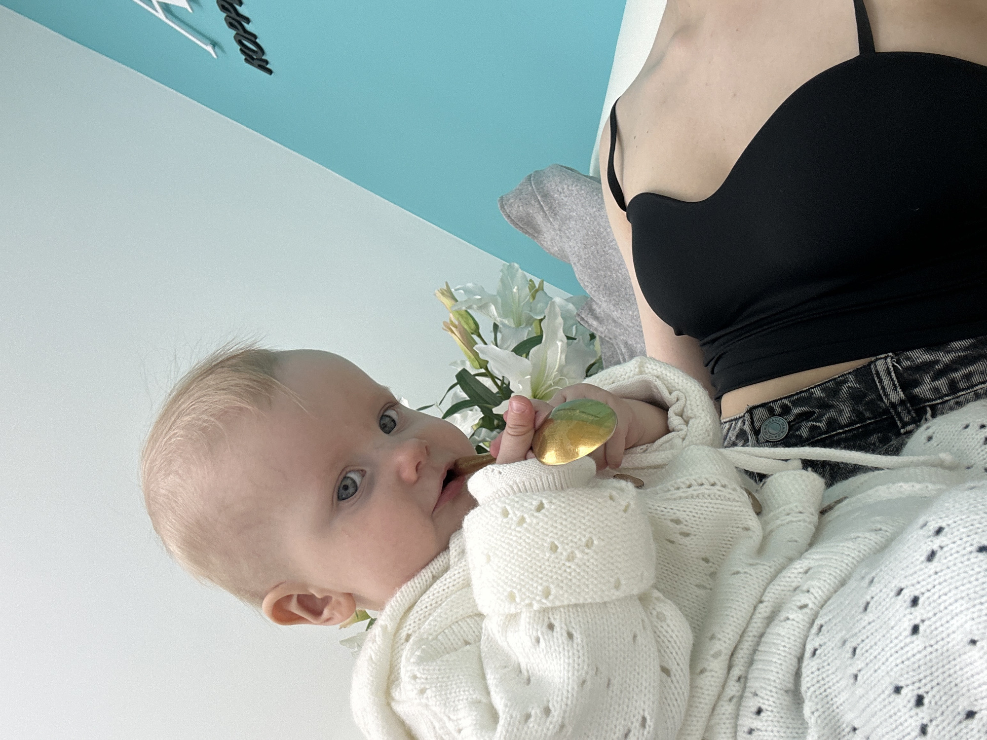 The image captures an indoor scene featuring an infant and a partially visible adult within a modern environment. A light-skinned baby with blonde hair and blue eyes, dressed in a white knitted cardigan featuring eyelet patterns, occupies the upper-left foreground, gazing directly at the viewer. The infant holds a reflective, metallic gold, tear-drop shaped object near its mouth, appearing to chew or gum it. To the right and slightly behind the baby, a bouquet of white lilies with green stems and leaves is present.
Below and to the right of the baby, the upper torso and midsection of an adult are visible, lying on a flat white surface. The adult wears a black strapless top or bra and dark, possibly denim, pants with a drawstring and a visible button. A grey fabric, potentially a blanket or garment, covers part of the adult's lower body.
The background wall is distinctly divided diagonally: the upper portion is a light blue-white, while the lower right section is a vibrant turquoise-teal. On this turquoise wall section, dark, three-dimensional letters are partially visible, spelling "HOPP", which is identifiable as part of "HOPPY". Based on this signage and the context of the subjects, the scene is located within a Hoppy Kids pediatric clinic or rehabilitation center in Moscow, Russia.