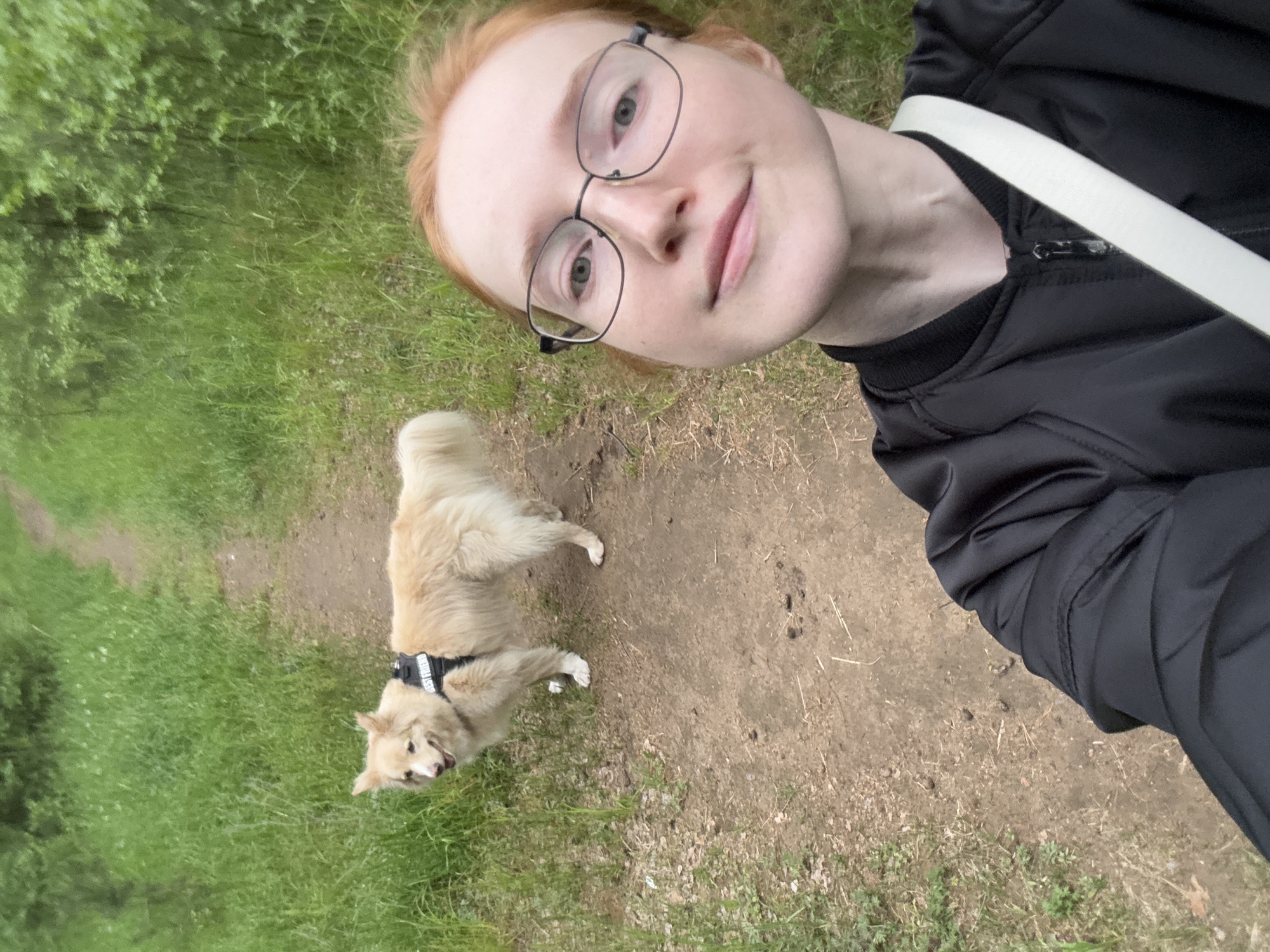 A young, light-skinned woman with red hair and glasses takes a selfie while walking her medium-sized, light-colored dog on a path. The path, composed of dirt and sparse vegetation, is bordered by green grass on the left and leads into a wooded area. The woman is wearing a black jacket with a white strap across her body. The dog wears a black harness. The location is an outdoor path, likely in a park, in the city of Chaikovsky, Russia.