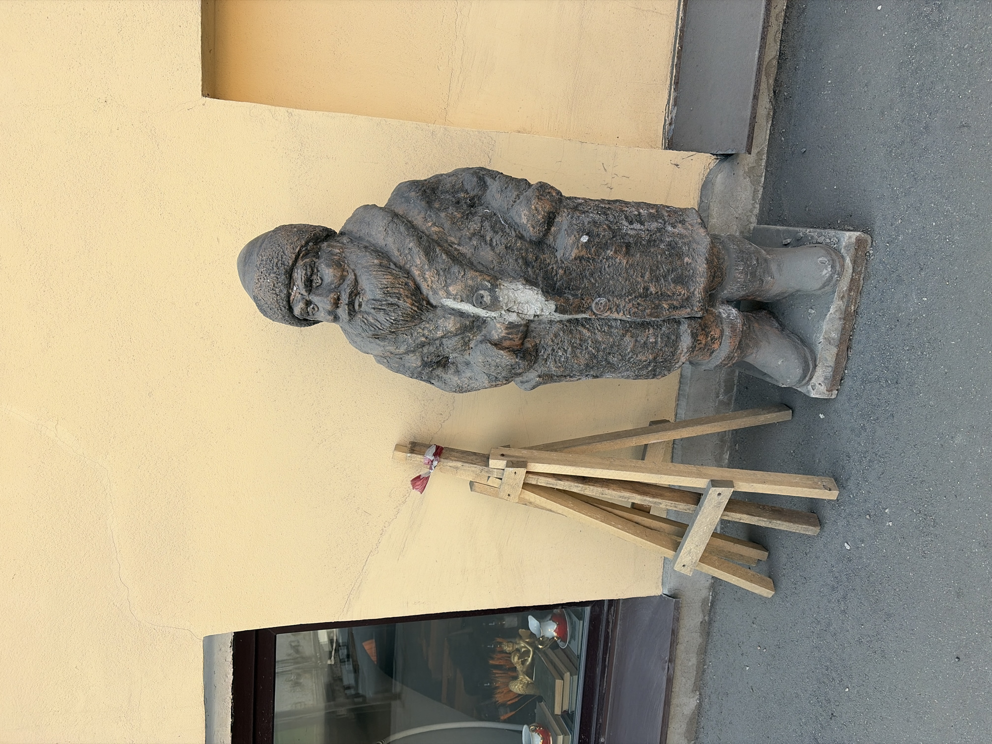 The image depicts a weathered, stone-like statue of an elderly man, likely a dwarf or gnome, standing on a plinth outside a building with a light yellow facade. The figure wears a cap and a long coat, and has a beard. Near the statue is a wooden easel leaning against the wall. Below is a window showing an interior display of decorative items. The scene is located in Saint Petersburg, Russia.