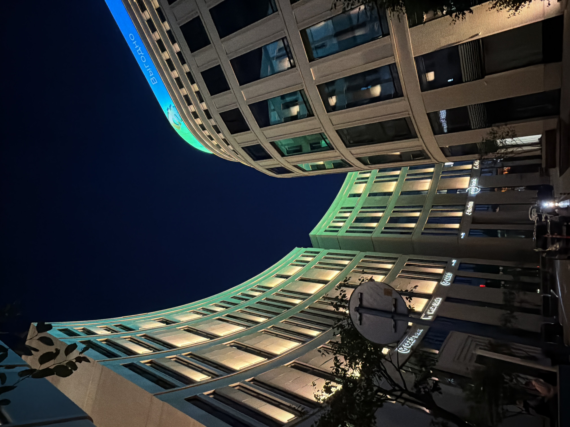 This is a low-angle, nighttime photograph of modern, curved buildings in Minsk, Belarus. The buildings are illuminated with a combination of green and yellow lights, highlighting their architectural features and numerous windows. One building prominently displays the word "Выгодно" in blue LED lights across its upper facade. The sky is a deep, dark blue. In the foreground, a street sign is visible, and the bottom of the image shows parked cars and some greenery. The overall scene conveys a sense of urban nighttime ambiance.