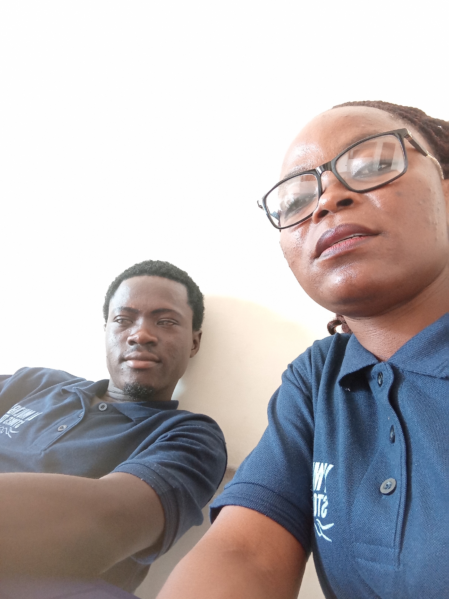 In Jalingo, Nigeria, a man and a woman are captured in a close-up, head-and-shoulders selfie against a plain, light-colored wall. The man, positioned on the left, is looking slightly off-camera with his arms crossed, wearing a dark blue polo shirt. The woman, on the right, is looking upwards, also wearing a dark blue polo shirt and black-rimmed glasses. The lighting is bright and appears to be natural, creating some glare on the woman's forehead and nose. The composition is casual, suggesting a candid moment.