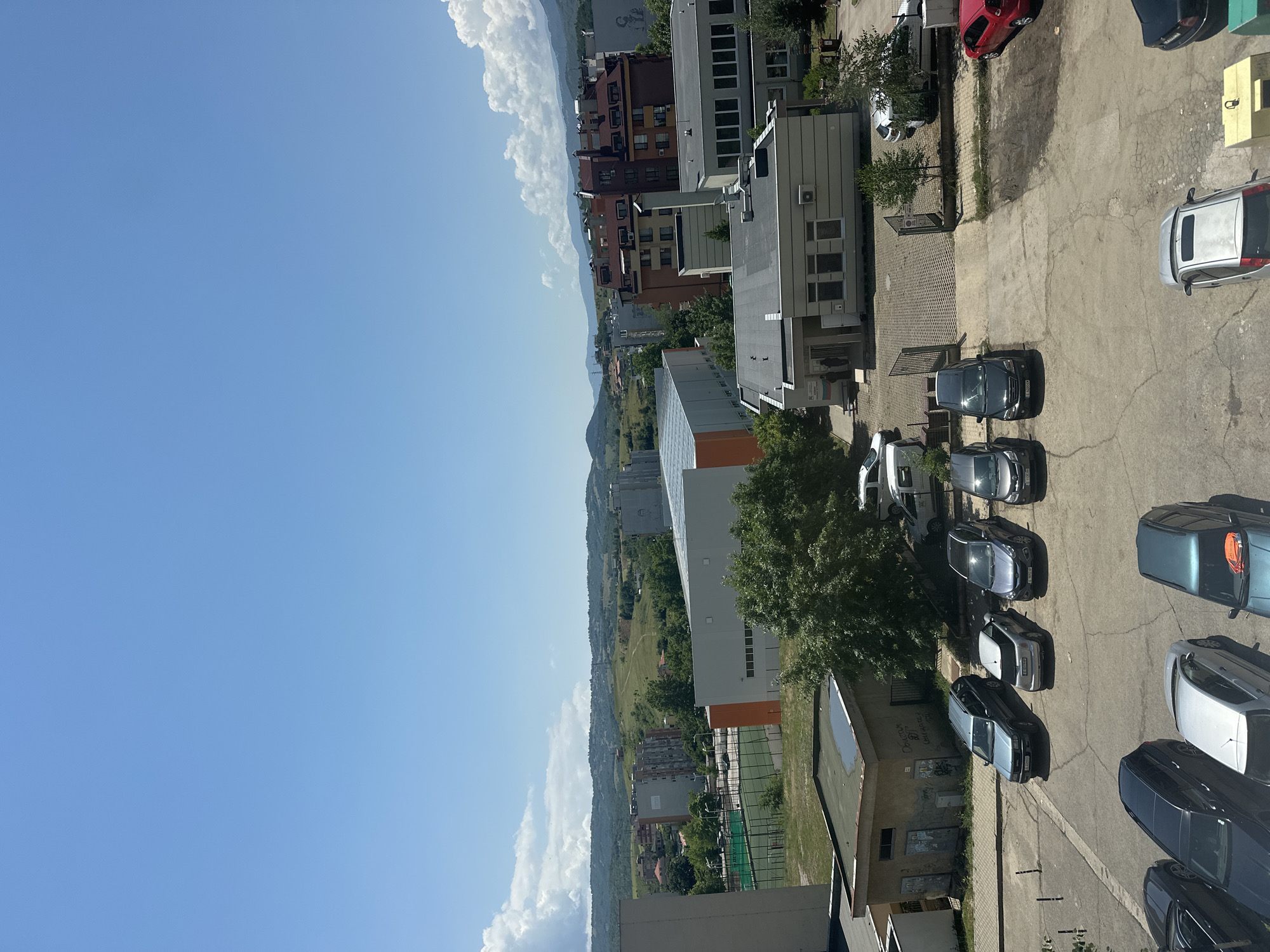 The image provides an elevated view of an urban landscape in Blagoevgrad, Bulgaria, under a clear blue sky with scattered white cumulus clouds.
In the foreground, a concrete parking lot is filled with numerous parked cars of various models and colors, including silver, black, dark blue, and red. The ground of the parking lot shows cracks and wear. Adjacent to the parking lot are parts of buildings, one of which is light-colored with visible windows and an air conditioning unit. Another building wall features Cyrillic graffiti, where the word "Обичам" (meaning "I love") is partially discernible. Sparse trees are interspersed around the parking area and between structures.
The midground reveals a denser concentration of buildings, ranging from multi-story residential blocks to more modern commercial structures, one notable for an orange strip along its side. A green sports court, possibly for tennis or basketball, is visible near these buildings. Beyond the immediate urban development, green fields and patches of trees stretch towards the horizon.
In the background, a range of hills or mountains is visible, partially obscured by the distant sky and clouds. The overall scene depicts a typical daytime environment in a city, with bright sunlight casting shadows from buildings and cars. No specific human activity or event is clearly taking place, with the scene primarily showcasing the static elements of the city.