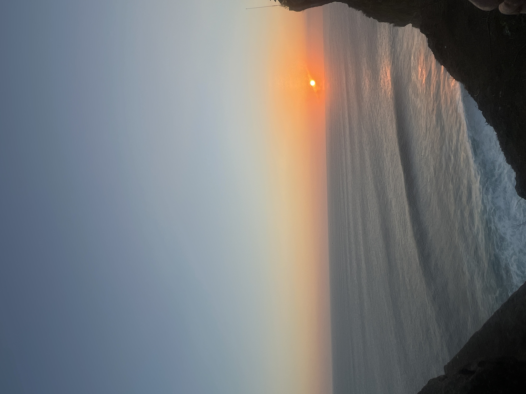 The image depicts a serene sunset over the ocean, viewed from a cliffside in Kuta Selatan, Indonesia. The sky transitions from a pale blue at the top to a vibrant orange and pink gradient near the horizon where the sun is setting. The sun itself appears as a bright, concentrated orb of light just above the waterline. Gentle waves with white foam crests are visible in the ocean, reflecting the warm hues of the sunset. A dark, rocky cliff face occupies the right side of the frame, with a thin, tall pole-like structure barely visible at its peak. The left side of the image shows another dark cliff edge, framing the expansive ocean view. The overall scene evokes a sense of tranquility and natural beauty.