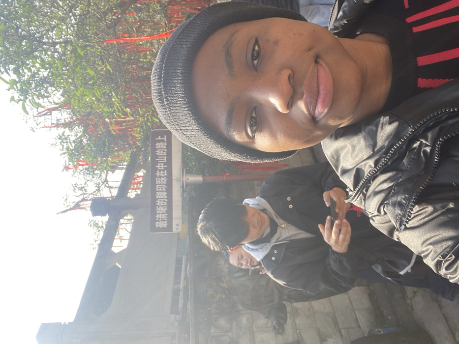A person wearing a beanie is taking a selfie outdoors. Behind them is another person looking at a phone, and a third person is partially visible. There are trees with red ribbons tied to them and a sign with Chinese characters. The area appears to be a park or garden on a sunny day. The picture is taken from a low angle, giving a perspective of looking up at the subject.