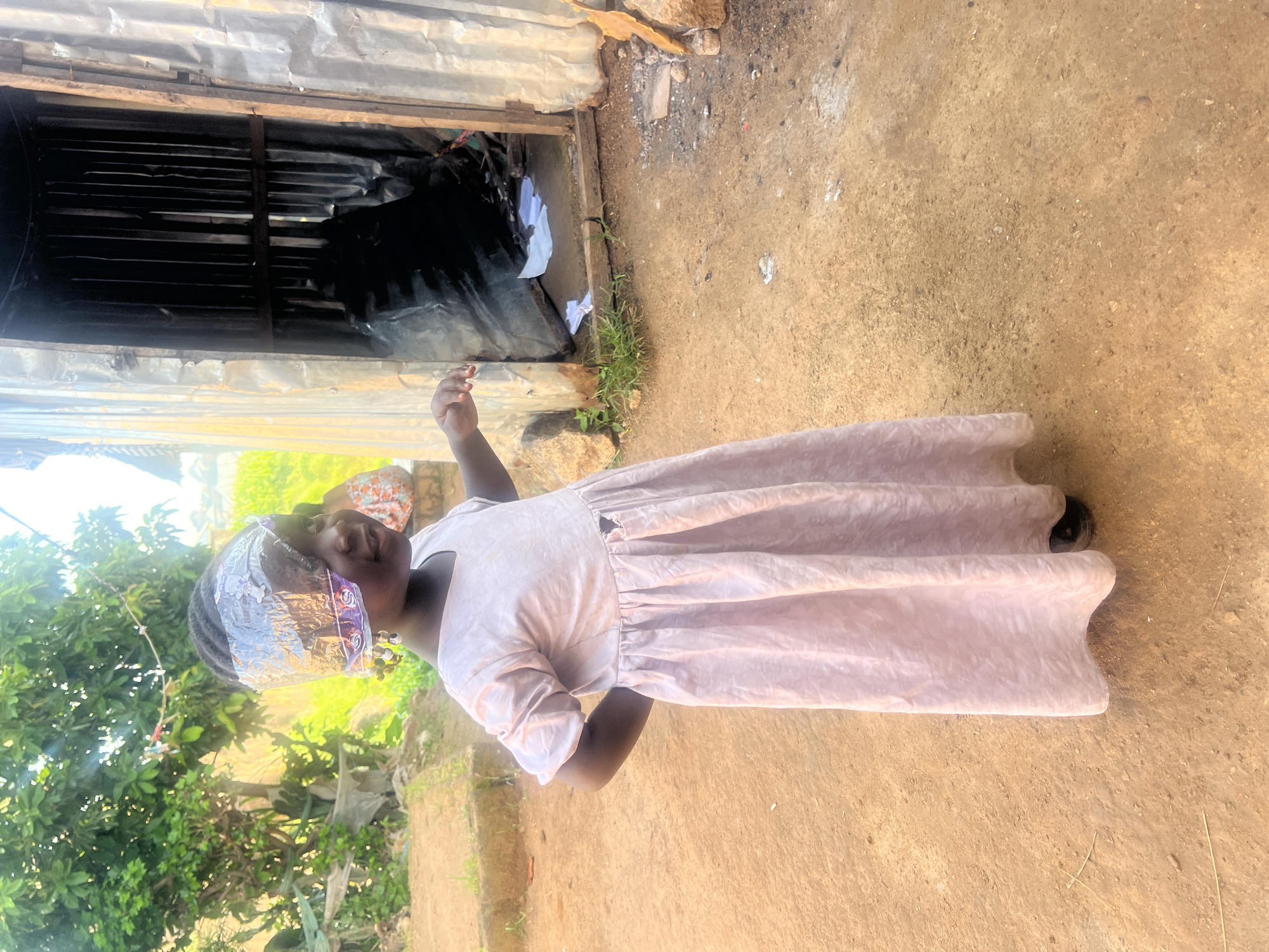 A young girl stands in front of a rustic structure in Jos, Nigeria. She wears a pale pink dress and has a piece of clear plastic wrapped around her head, adorned with colorful baubles as earrings. Her hands are placed on her hips, and she is looking to her right with a slight smile. The structure behind her is made of corrugated metal and weathered wood, with an open doorway revealing a dark interior. The ground is dusty earth, with scattered stones and patches of grass. The background shows trees and foliage under a bright sky.