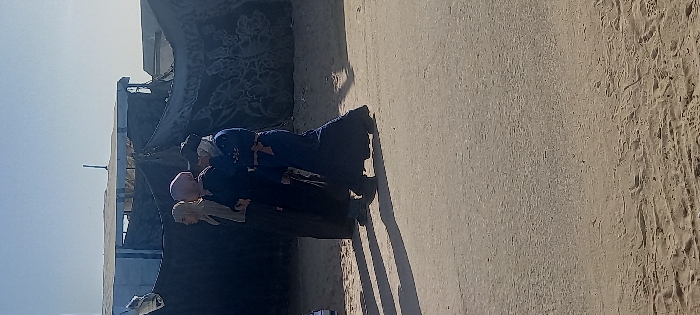 An outdoor, daylight scene in Khan Yunis, Palestinian Territory, depicts three individuals, appearing female, walking from left to right across a dirt road. The person on the left wears a light beige head covering and a dark, long dress. The middle person wears a light pink head covering and a dark long dress. The person on the right wears a dark head covering, a dark blue long dress with light embroidery on the upper torso, and a tan belt or sash. Strong shadows from the individuals are cast to their left on the unpaved, light brown ground, which shows tire tracks in the foreground. In the background, a light blue structure, possibly corrugated metal or painted wood, is visible above a dark tarpaulin or fabric screen displaying light-colored patterns. The sky is clear and light blue.