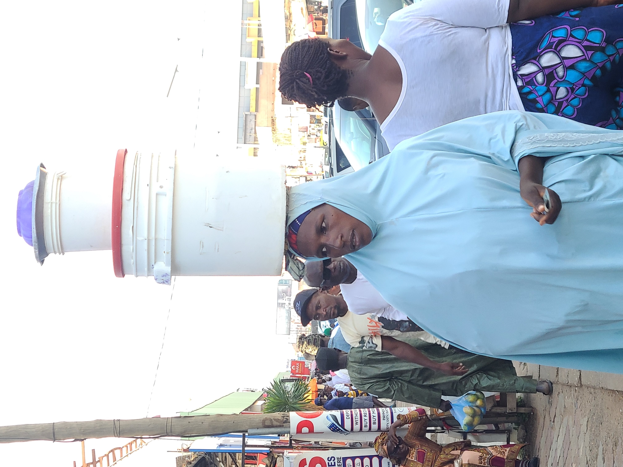 This is a medium-shot, eye-level, outdoor photograph taken in Jos, Nigeria. In the foreground, a woman wearing a light blue hijab and a matching gown faces slightly to the left, carrying a stack of white buckets on her head, topped with a purple lid. Her gaze is directed forward. Behind her, a woman in a white t-shirt and a patterned skirt is partially visible from the back. The background shows a street scene with other pedestrians, including a man in a green robe carrying a bag of fruit, and a man in a light-colored t-shirt wearing a baseball cap. Several vehicles are parked on the street, and buildings line the background, suggesting a busy urban environment. The overall lighting indicates it is daytime.