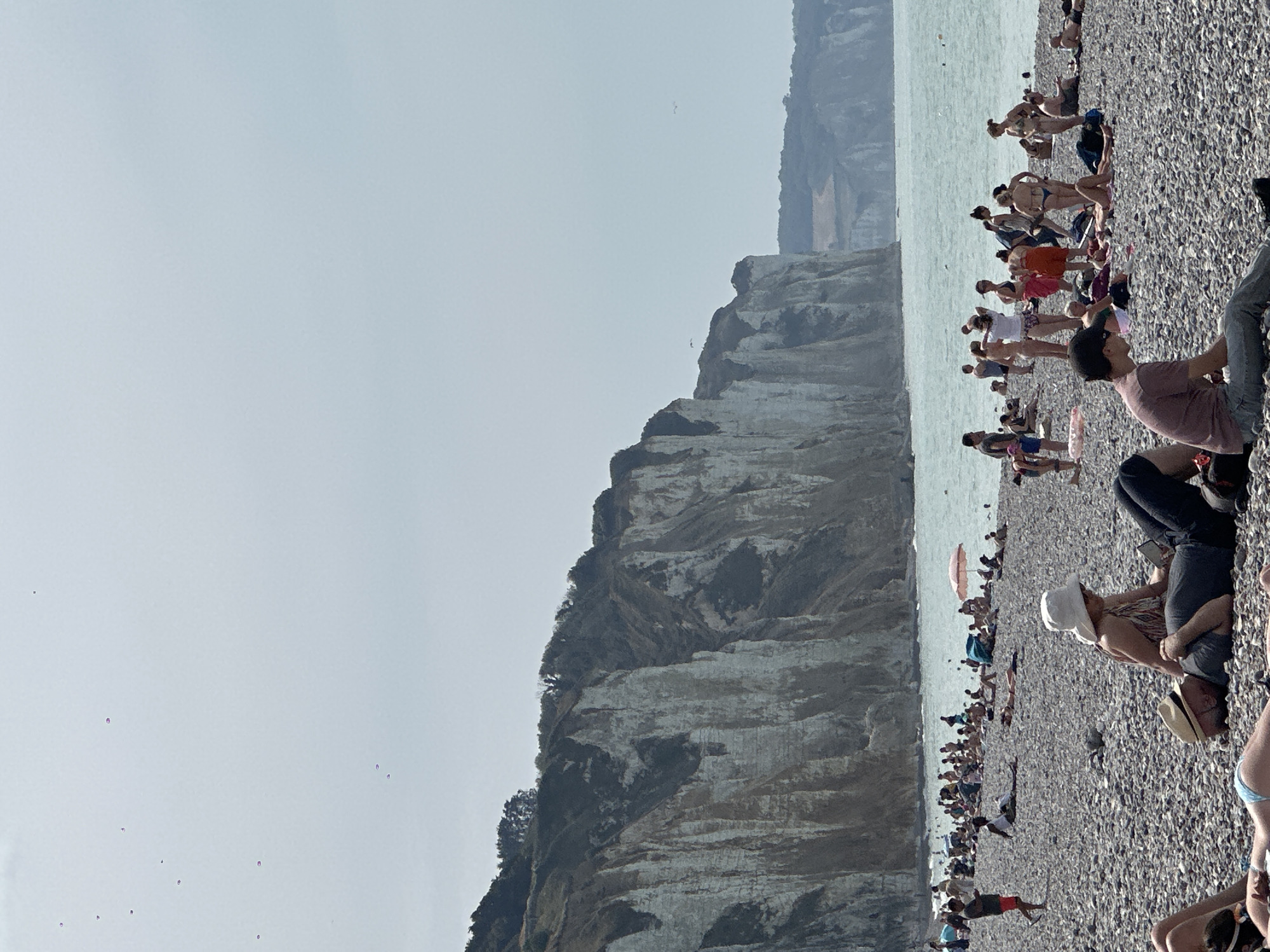 This is a photograph taken at the pebble beach in Dieppe, France, showcasing a bustling scene on a sunny day. The background is dominated by imposing white chalk cliffs, characteristic of the region, with a hazy sky overhead. The sea is visible, calm and a pale blue-green. The beach is populated by numerous people enjoying the weather and seaside.
In the foreground, several individuals are relaxing on the pebbles. A man wearing a straw hat lies on his back, while a woman in a white sun hat sits beside him, looking at a phone. Further down, another person in a pink shirt is seated. To the left, a person wearing a blue bikini bottom is lying on their back.
In the midground, a large number of people are scattered across the beach. Some are sunbathing, while others are standing, talking, or wading in the shallow water. A small pink umbrella provides shade for a group. A child with a red ball is visible near the water's edge. Further back, more people are visible in clusters, and a few can be seen in the water.
The overall impression is one of a lively summer day at a popular tourist destination, with the dramatic cliffs providing a striking natural backdrop. The pebbles underfoot are varied in size and color, typical of a beach formed by erosion from the cliffs. The light suggests it is midday or early afternoon.