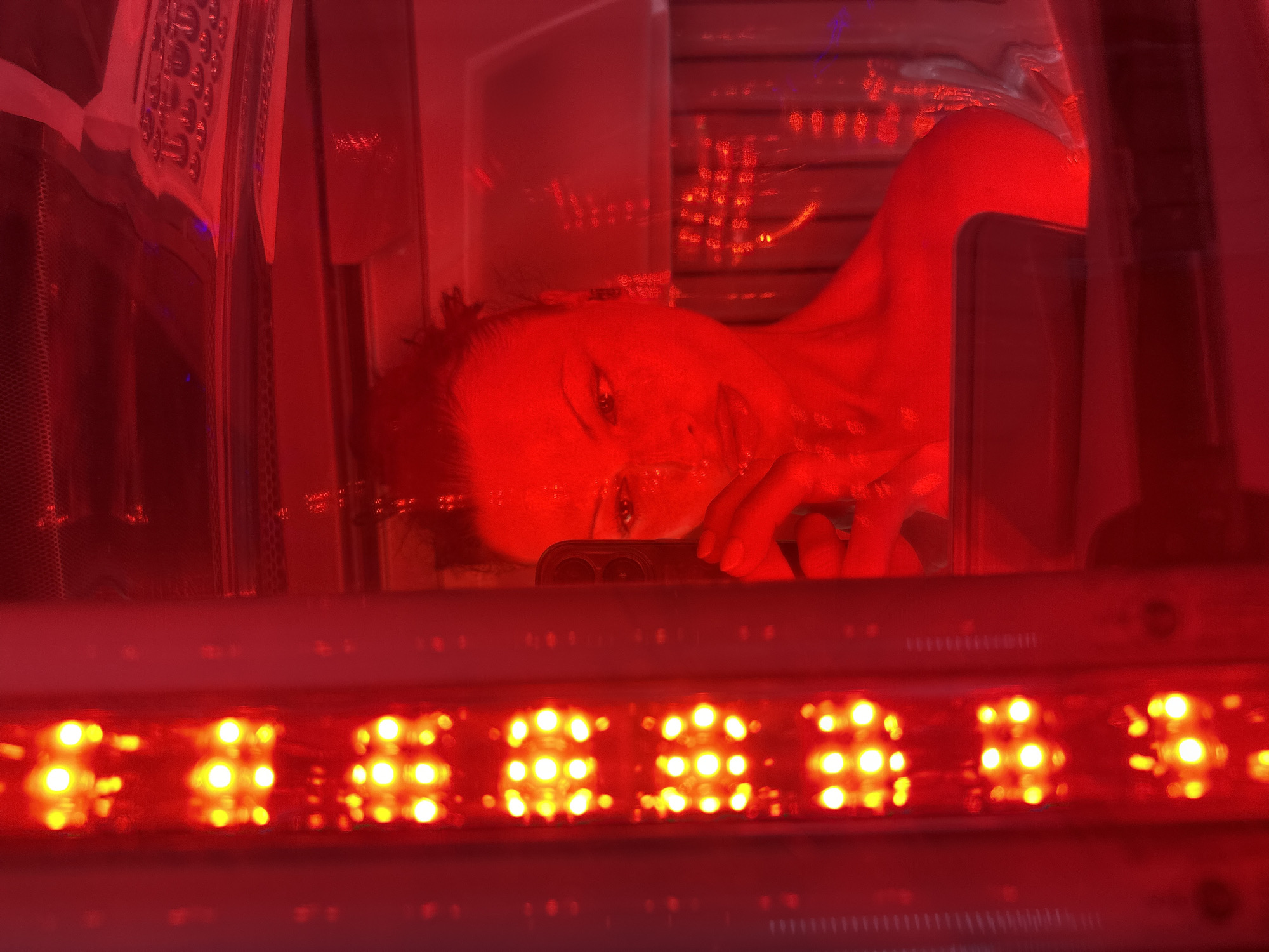 The image captures a solitary adult female inside an enclosed red light therapy or tanning booth, located in Tunbridge Wells, United Kingdom. The specific address within the city cannot be determined from the visual information. The individual, appearing to be an adult female with dark hair pulled back, is visible in reflection, bathed in an intense, uniform red glow emanating from numerous bright, circular red LED lights arranged vertically along internal panels. She is holding a dark-colored smartphone with a triple camera array in her right hand, positioned to take a self-portrait. Her face, partially visible, features prominent eye makeup and red lipstick; her expression is neutral. She appears to be shirtless, revealing bare shoulders and upper chest, consistent with participation in a beauty or wellness treatment. In the upper right background, a control panel with multiple circular buttons and a fine mesh grille are faintly discernible, indicating the functional components of the booth. The pervasive red illumination dominates the entire scene, casting a deep reddish tint over all surfaces and the individual's skin.