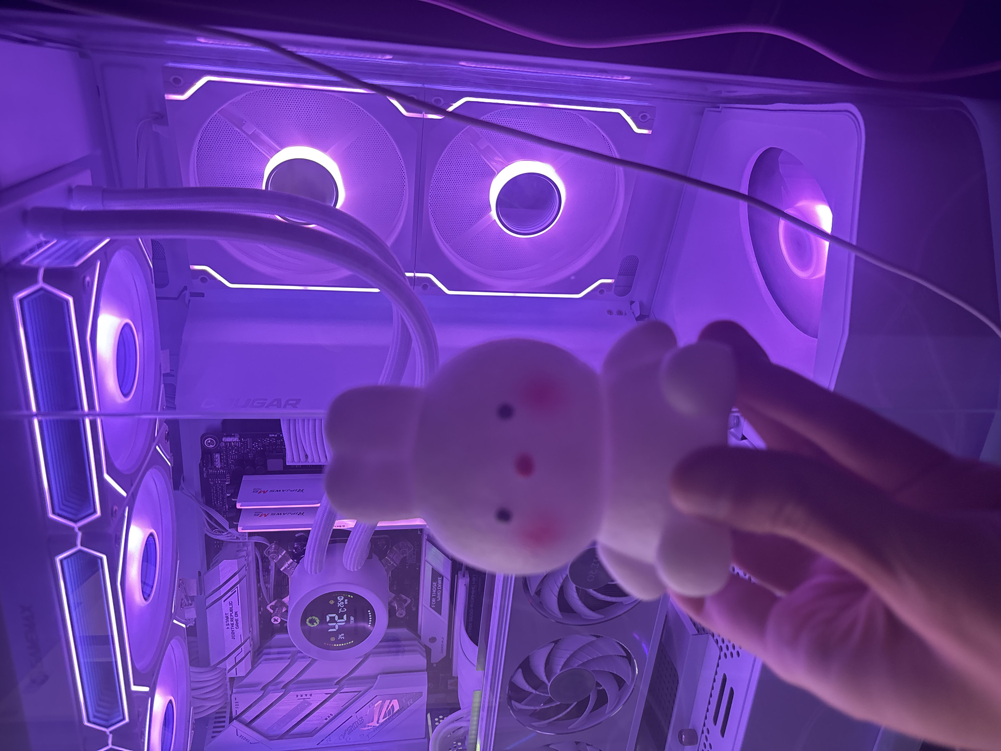 A hand holds a small, white, bunny-shaped figurine in front of a computer tower. The computer tower has purple LED lighting and visible internal components including fans, a motherboard, and a liquid cooler with a digital display showing "42" degrees Celsius. The background is dark, suggesting the image was taken in a dimly lit room. The location is Unknown, Unknown.